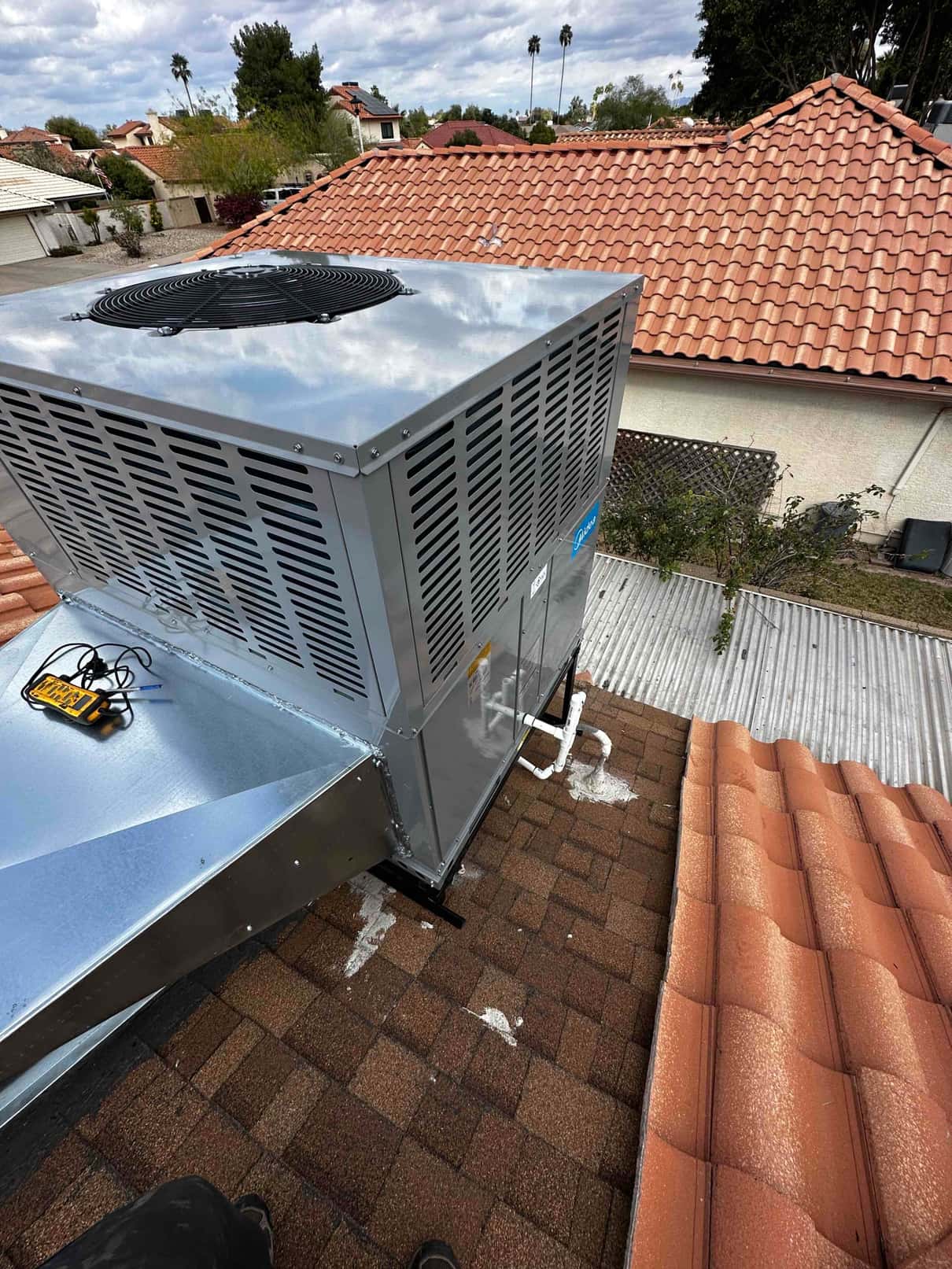 New rooftop packaged HVAC unit set on shingles with sealed curb, metal supply plenum, and condensate drain piping visible.