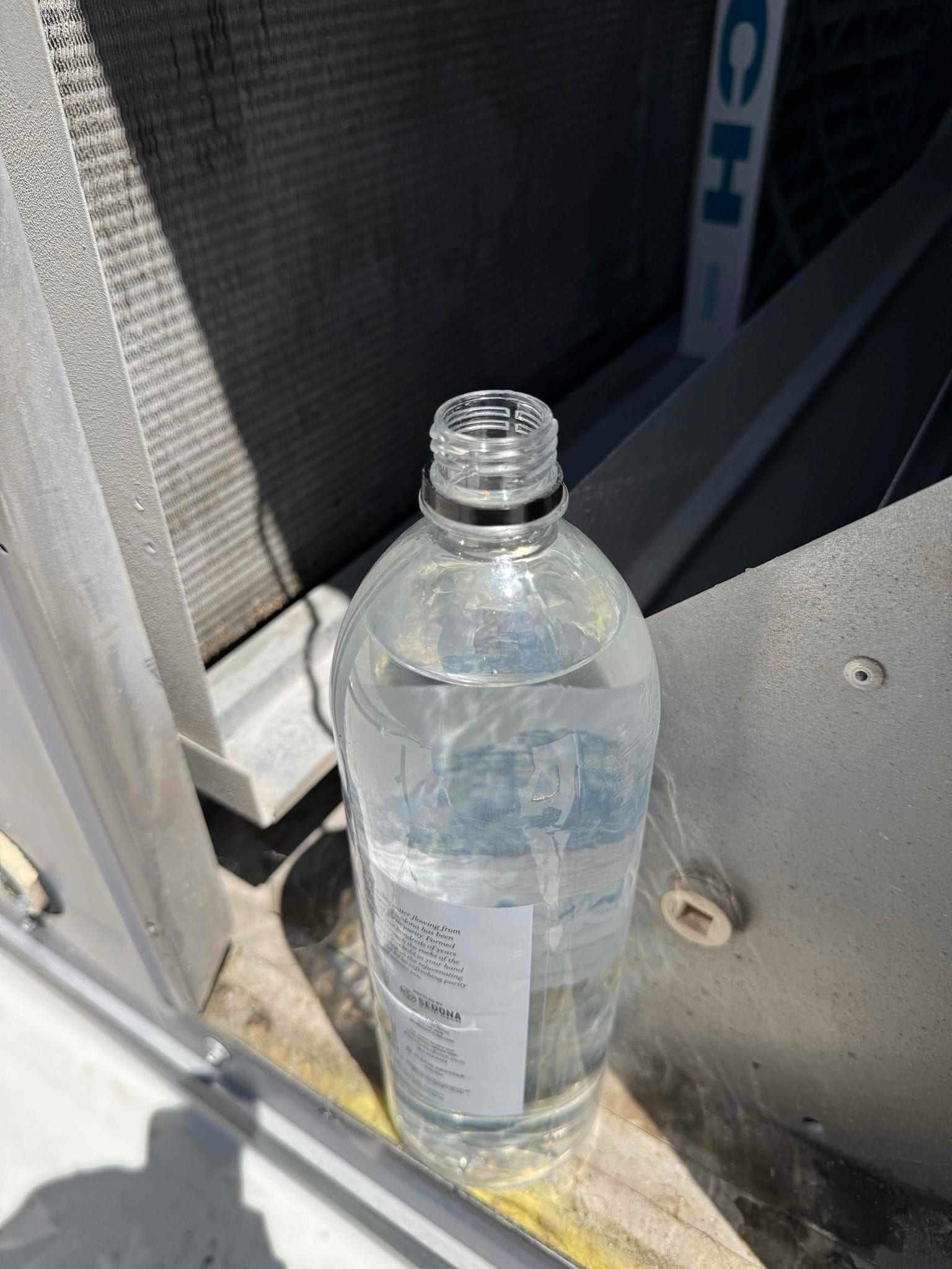 Open water bottle placed inside an outdoor HVAC condenser cabinet near the coil and fan shroud during maintenance.