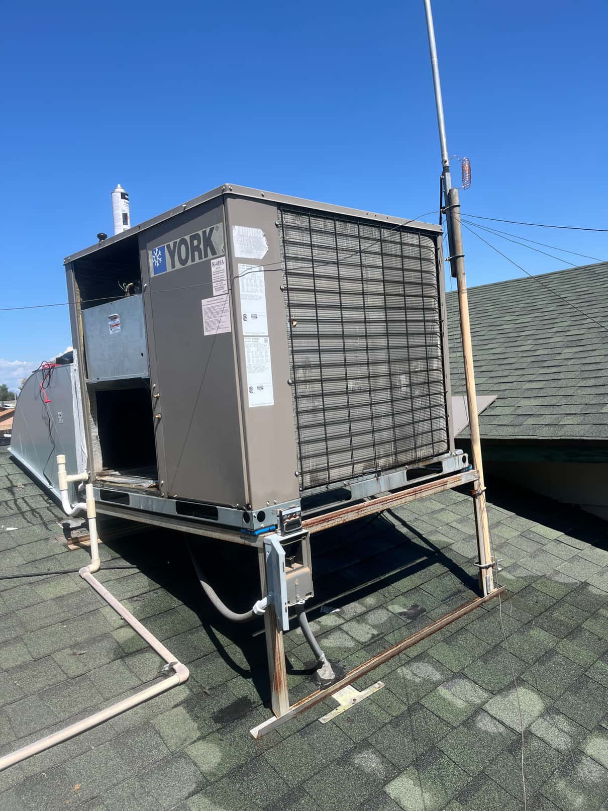 Rooftop packaged HVAC unit on a metal stand, showing condenser coil, service panels, condensate drain piping, and refrigerant lines.