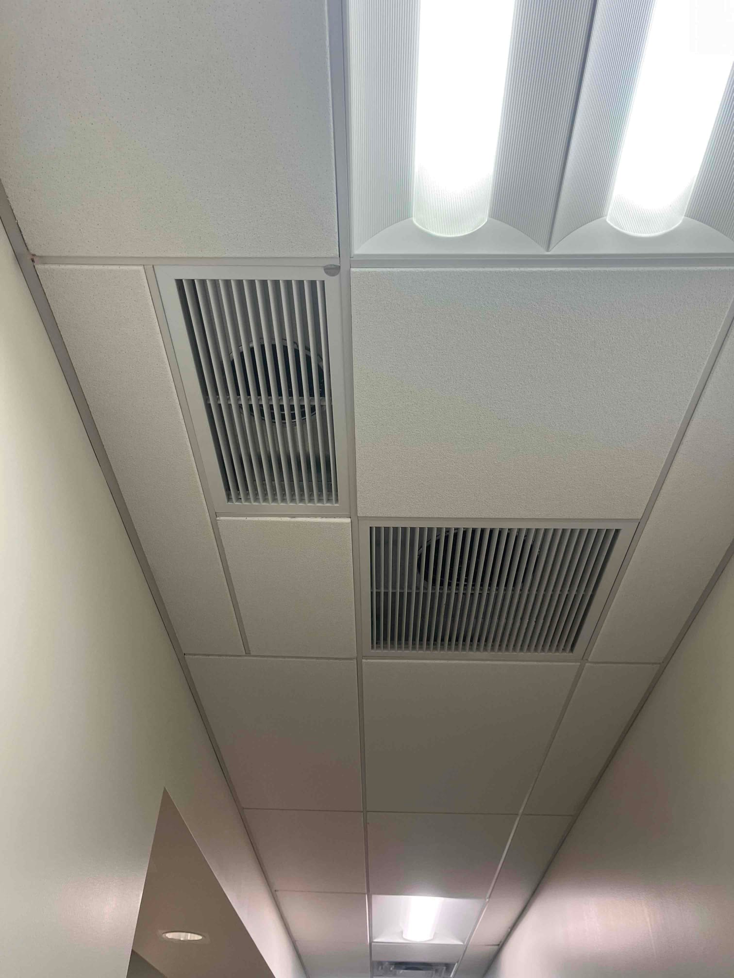 Suspended ceiling with fluorescent lights and two rectangular supply and return air grilles along a hallway.
