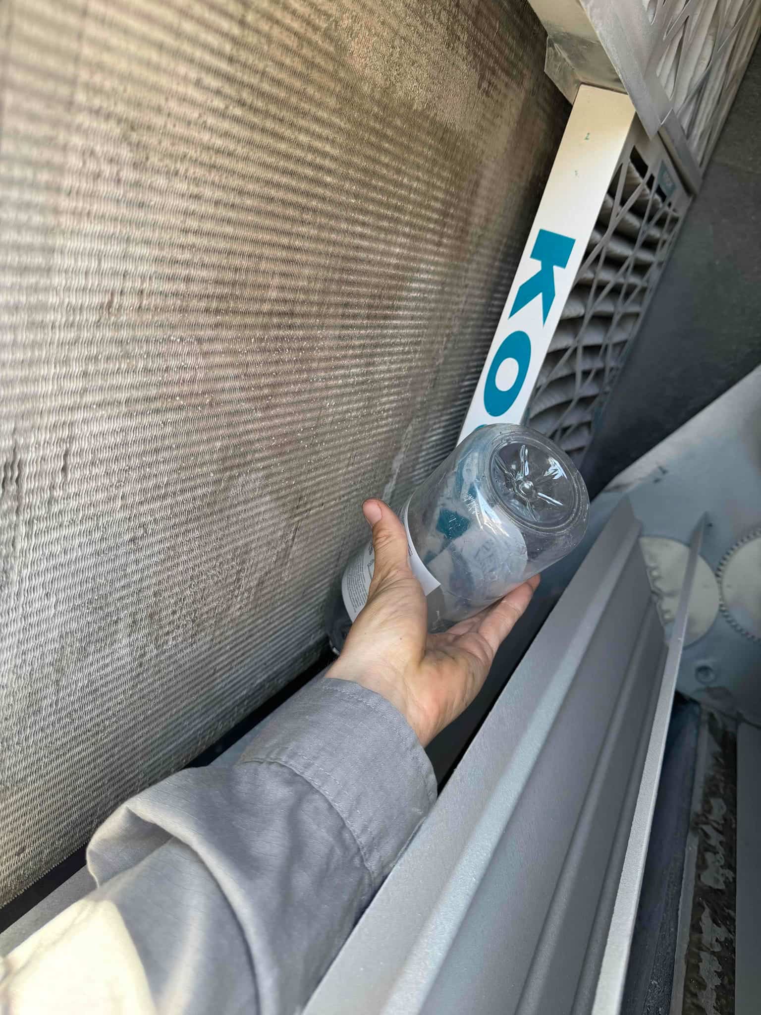 Technician’s hand holds a clear container next to a dirty evaporator coil and adjacent air filter housing.