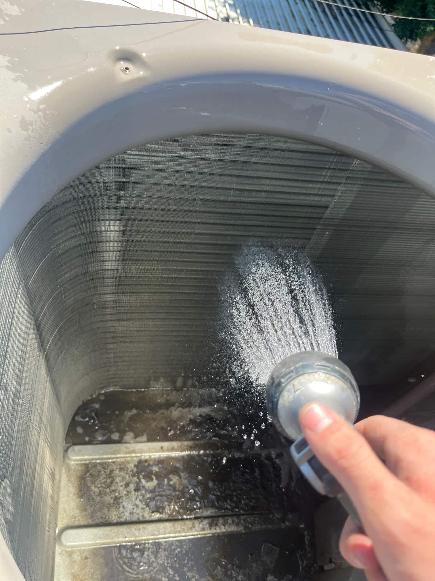 Technician rinses debris from condenser coil fins inside the outdoor unit housing during evaporator coil cleaning and AC maintenance.