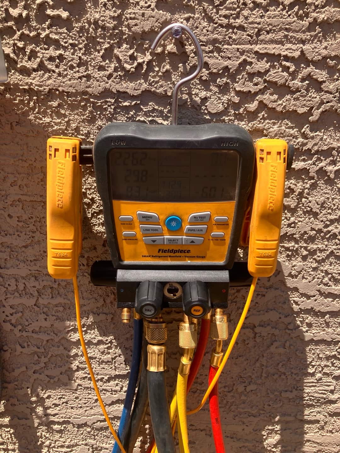 Digital manifold with temperature clamps connected to refrigerant hoses during commissioning work on an exterior stucco wall.
