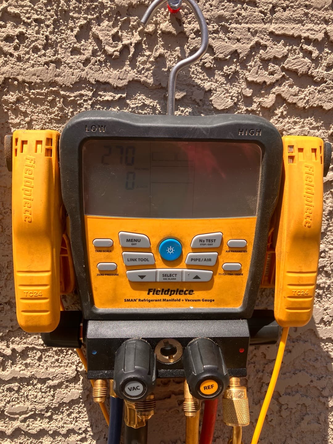 Digital manifold with thermocouple clamps attached hangs on a wall; screen shows partial readings but operating pressures are not clearly visible.