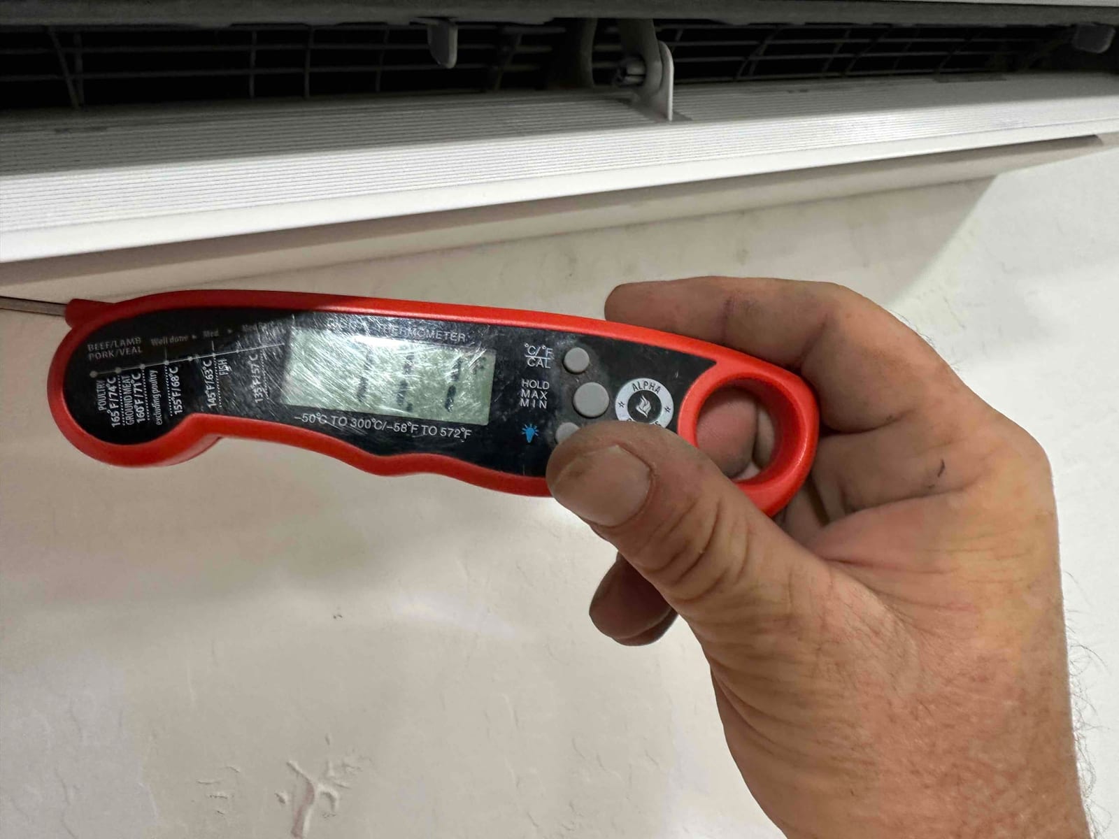 Digital probe thermometer held at a wall-mounted indoor unit outlet to measure supply air temperature during commissioning.