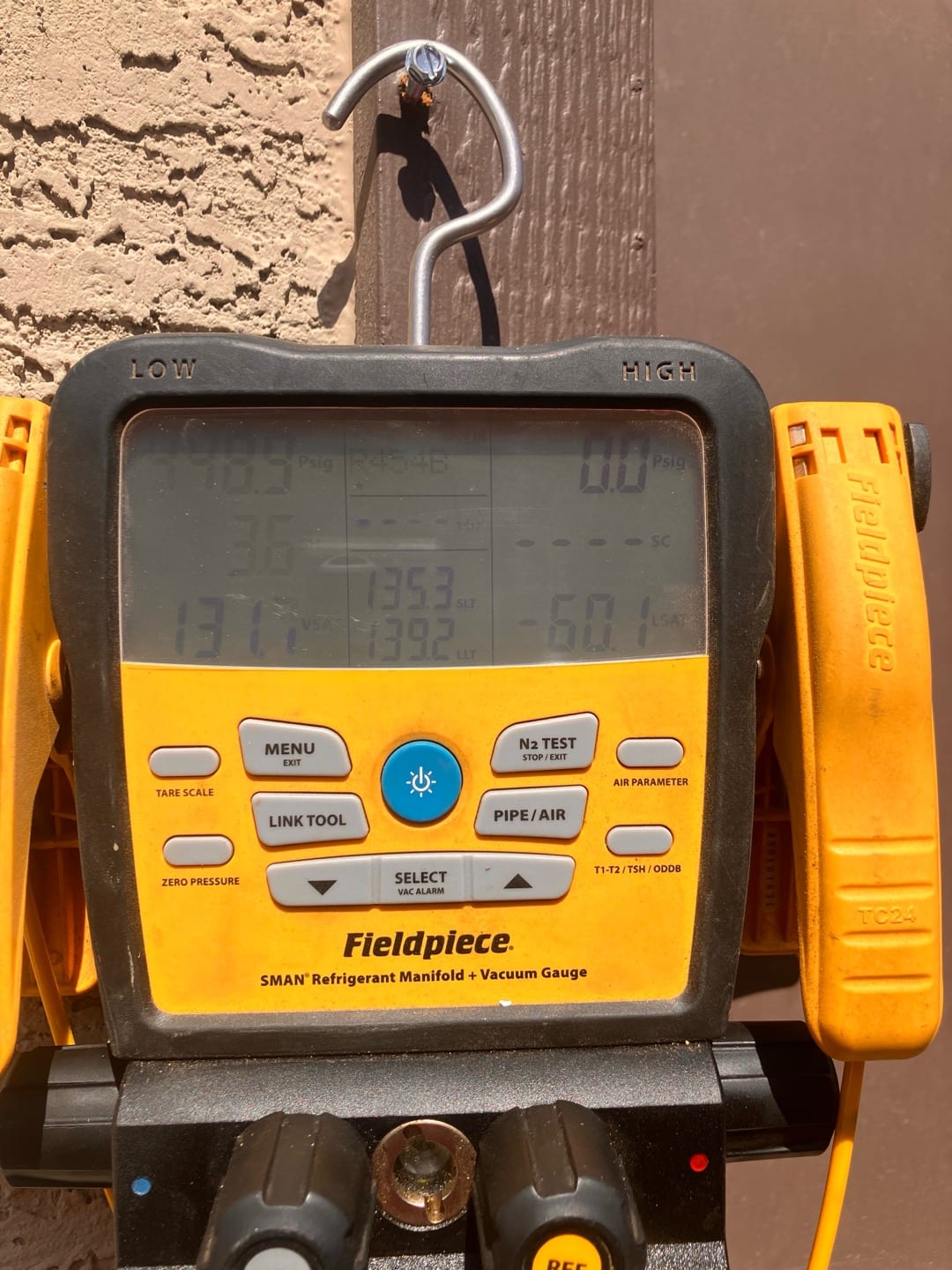 Digital refrigerant manifold and vacuum gauge hanging at the outdoor unit, screen sun-washed with unreadable values.