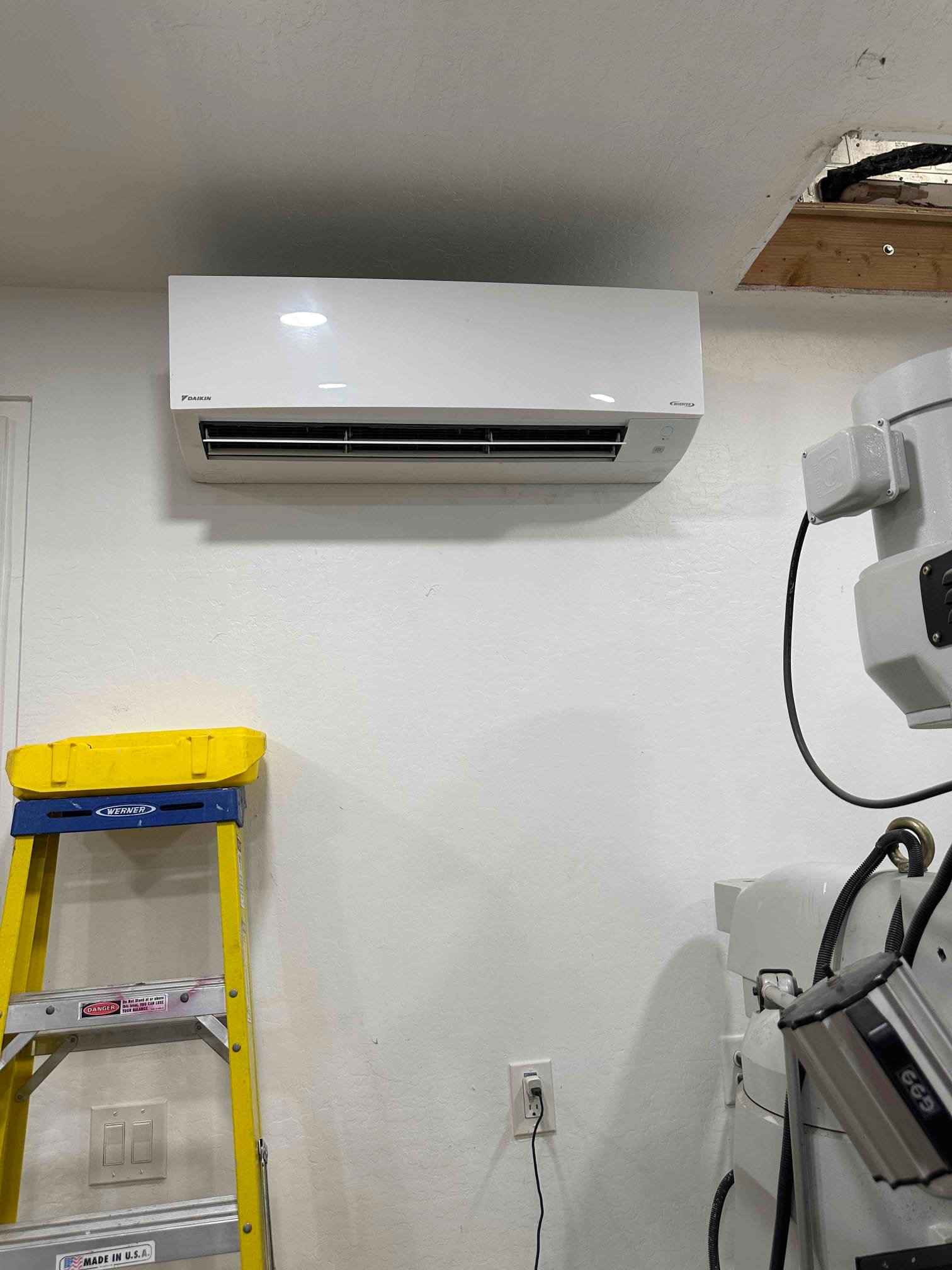 Wall-mounted mini-split indoor unit newly installed and powered, with nearby ladder and finished wall penetration visible.