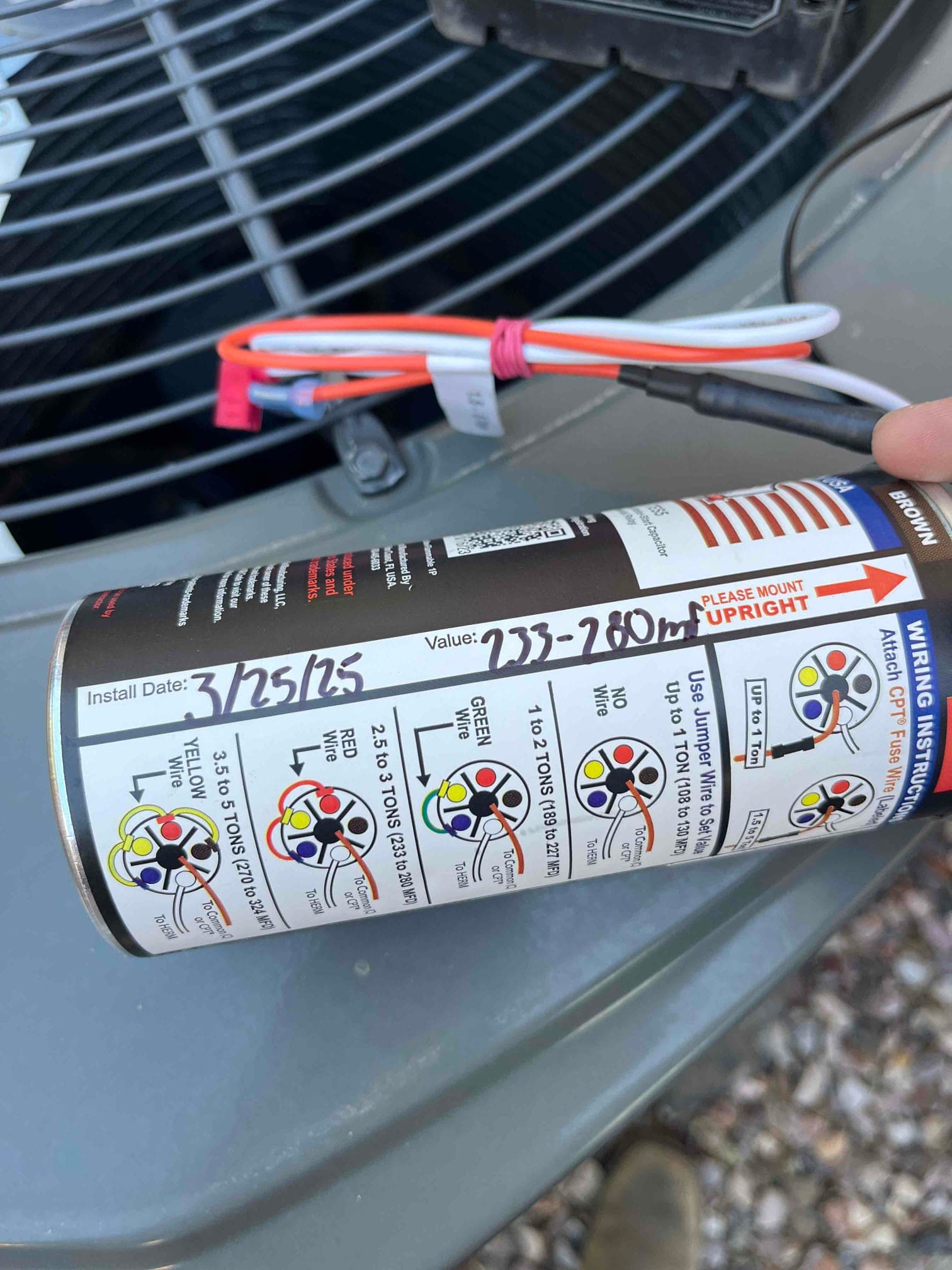 Cylindrical start-assist component with wiring instruction label, handwritten install date, and value marking, held beside outdoor condenser fan grille.