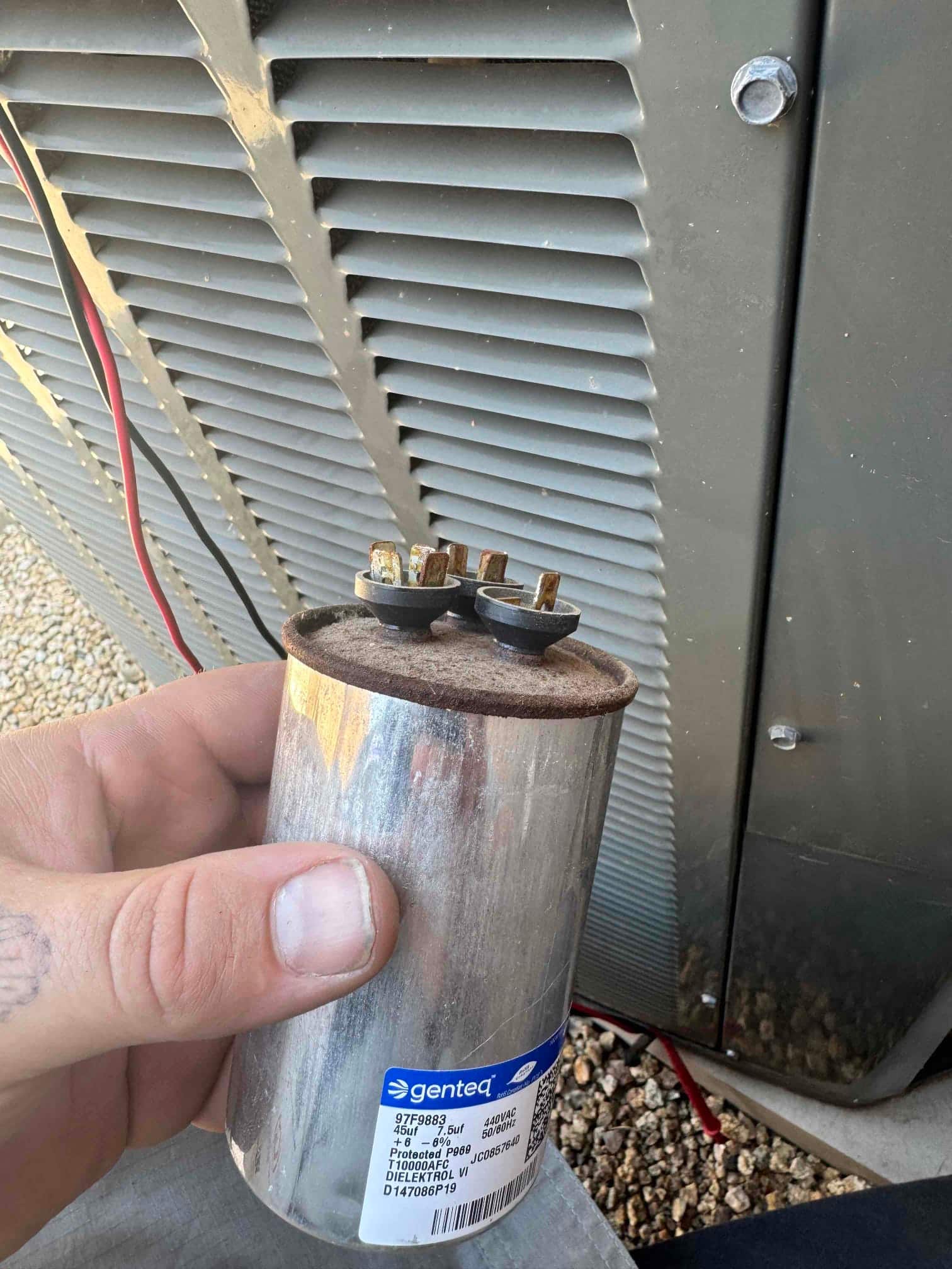 Dual-run capacitor labeled 45/5 μF, showing rust on the top cover and corroded spade terminals, held beside the outdoor condenser housing.