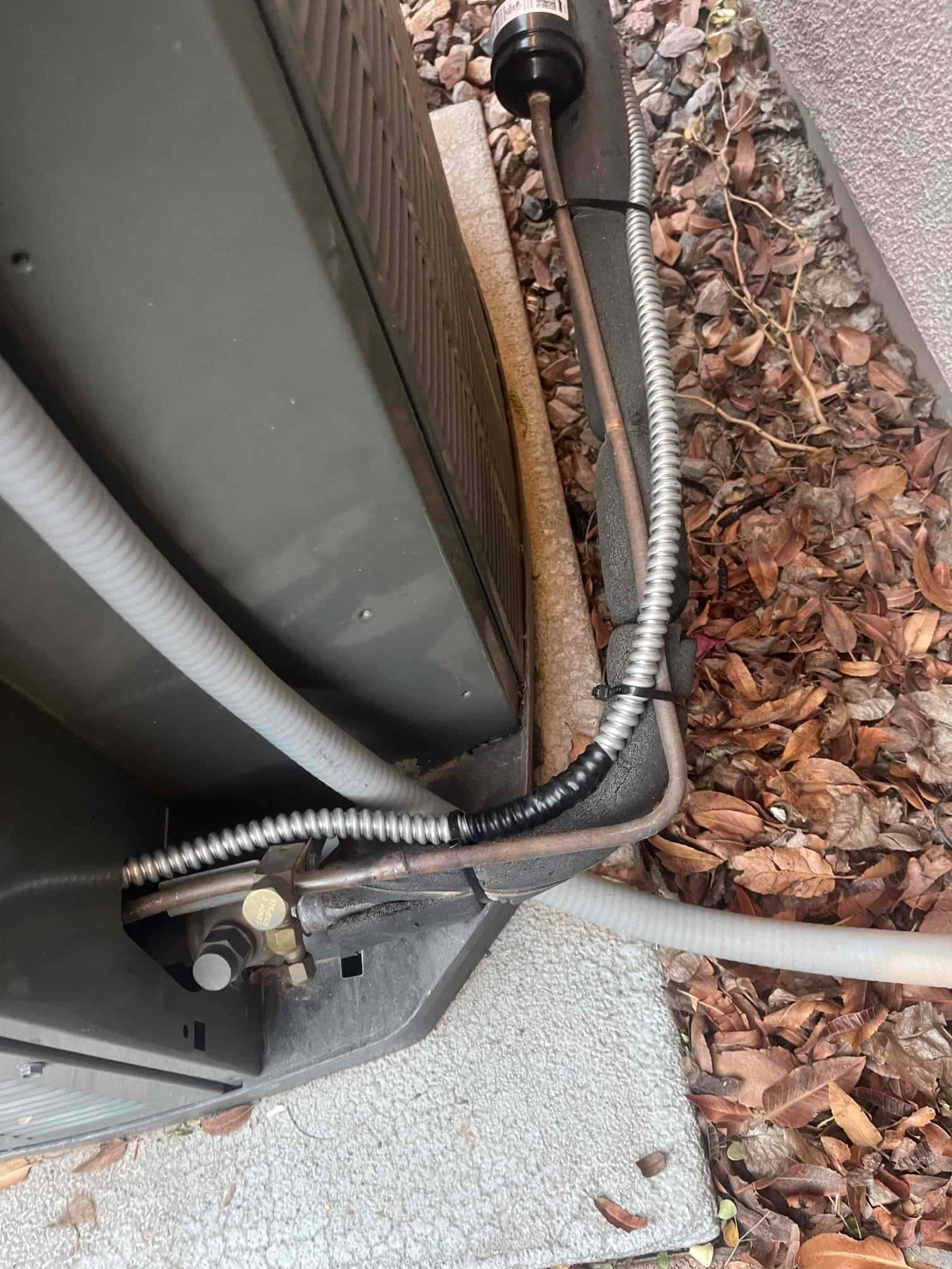 Outdoor condenser corner showing refrigerant service valves, insulated suction line, liquid line with inline filter-drier, and flexible electrical conduit.