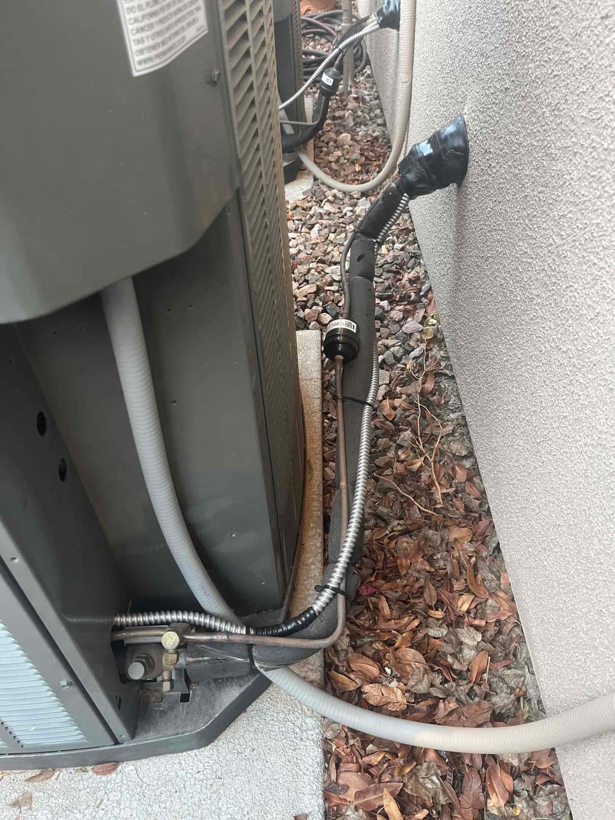 Outdoor condenser showing insulated refrigerant line set, service valves, and flexible electrical conduit entering the exterior wall.