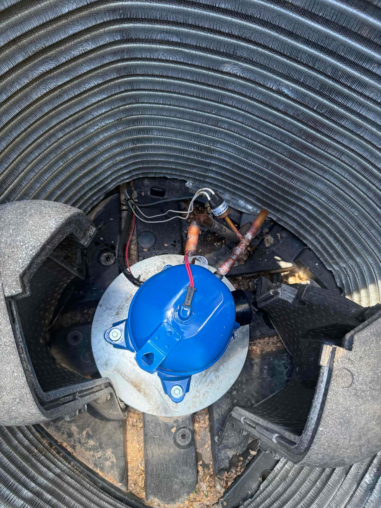 Outdoor condenser interior showing blue hermetic compressor with wiring and surrounding condenser coil and copper refrigerant lines.