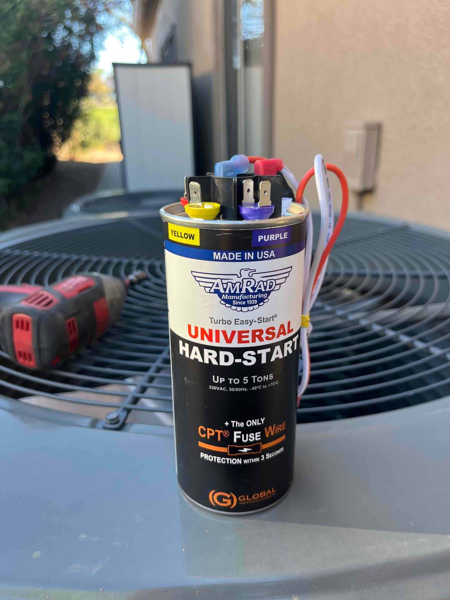 Universal hard-start capacitor kit with colored terminals and attached leads resting on an outdoor condenser top. Label states up to 5 tons.