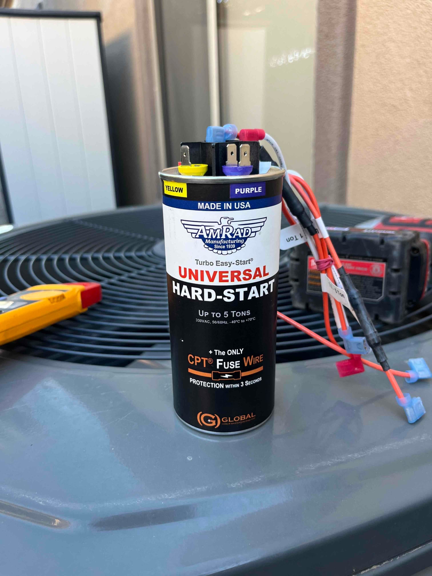 Universal hard-start capacitor with labeled terminals and attached wires sitting on outdoor condenser top.