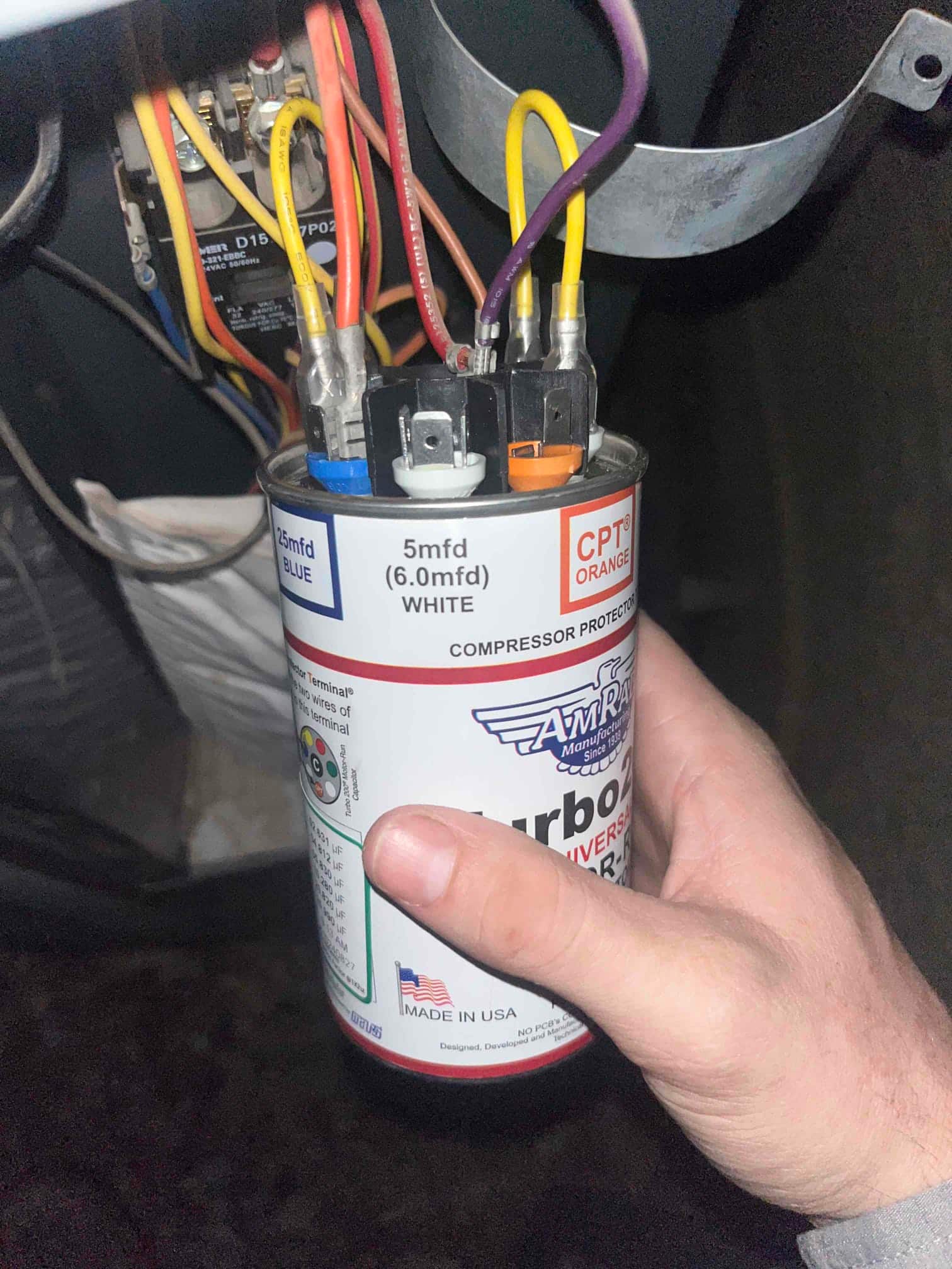 Universal run capacitor held by technician, showing color-coded terminals with connected wires and printed ratings 25 mfd BLUE and 5 mfd WHITE.