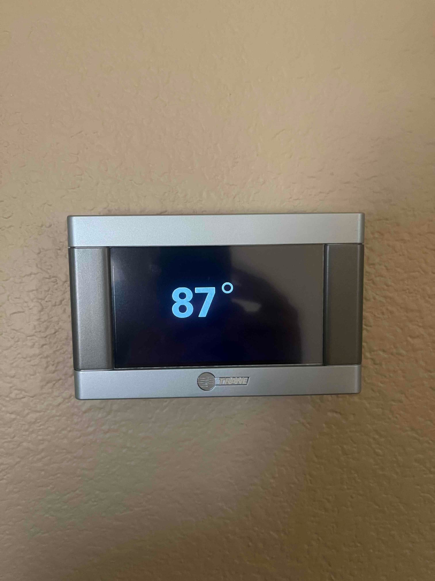 Wall-mounted thermostat with a black display shows 87° in large digits against the surrounding beige wall.