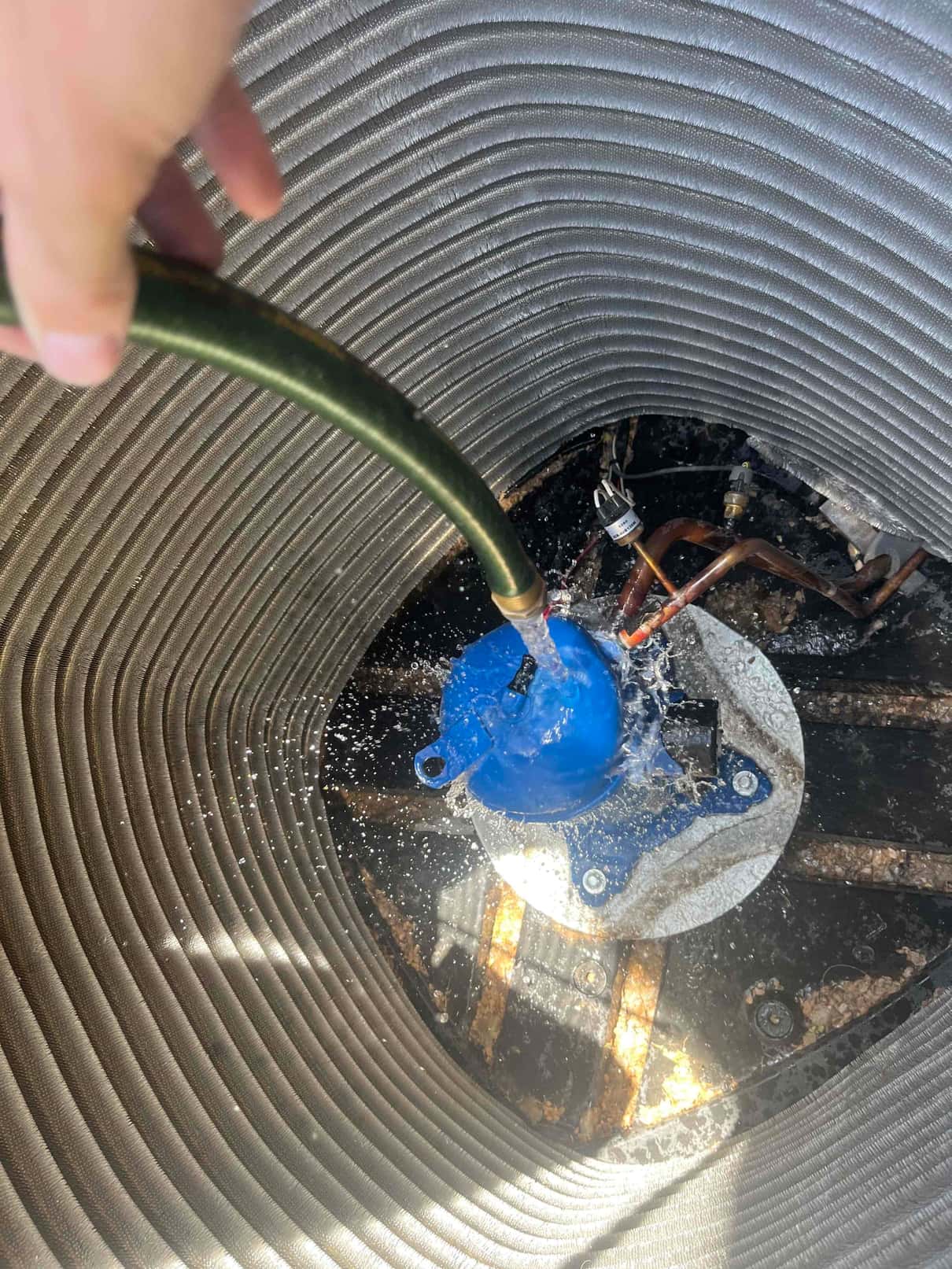 Water from a hose sprays onto the blue condenser fan motor and mounting plate inside the outdoor unit.