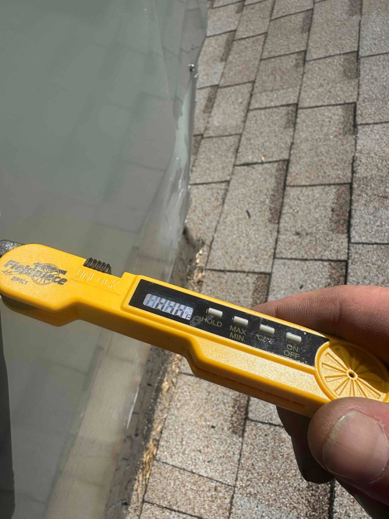 Digital thermometer-style meter held beside rooftop HVAC equipment; display is unreadable due to glare.