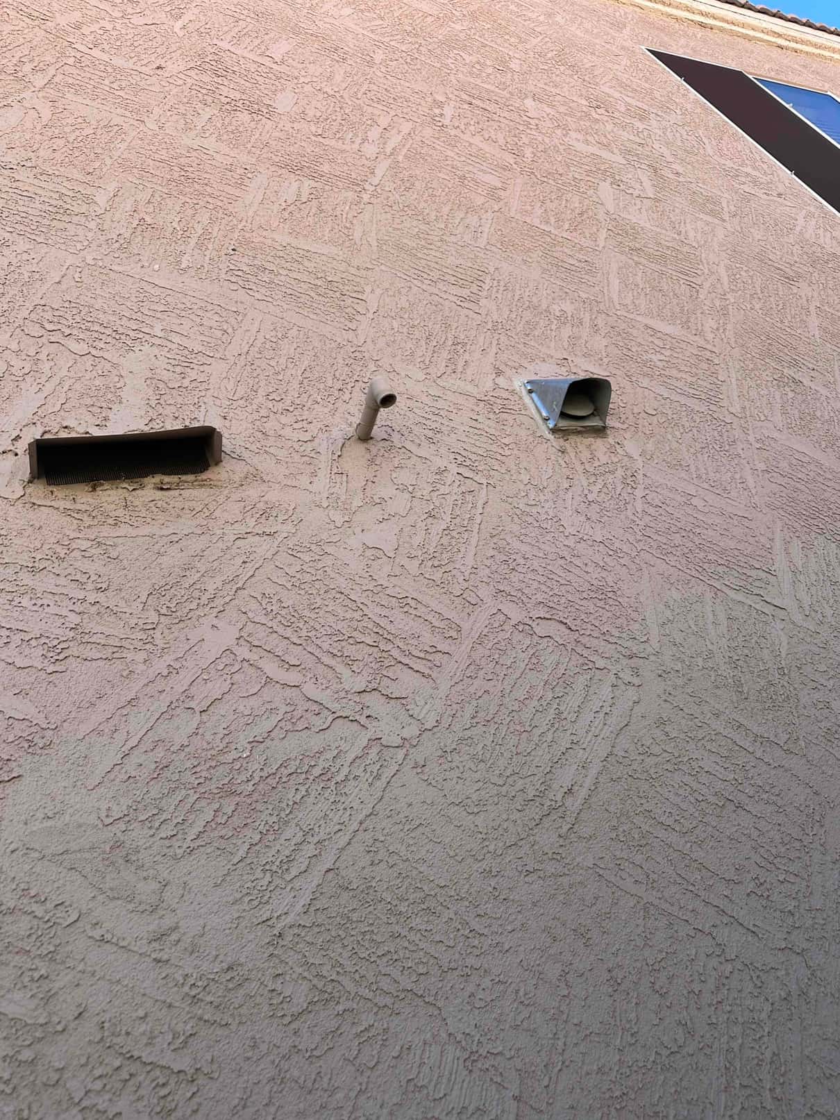 Exterior stucco wall shows a rectangular vent, a short PVC drain pipe, and a metal hooded vent.