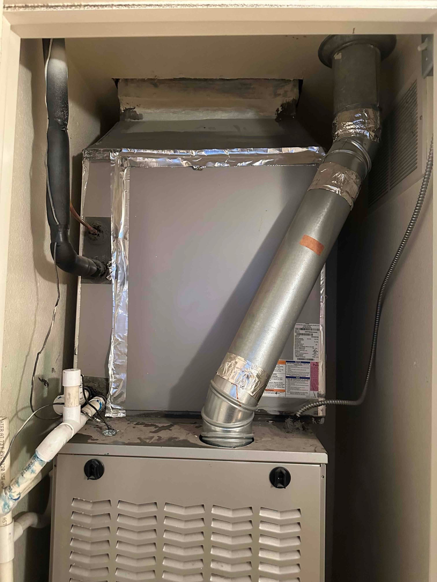 Indoor gas furnace and evaporator coil cabinet with foil-taped seams, insulated refrigerant line, condensate drain, and metal vent pipe visible.