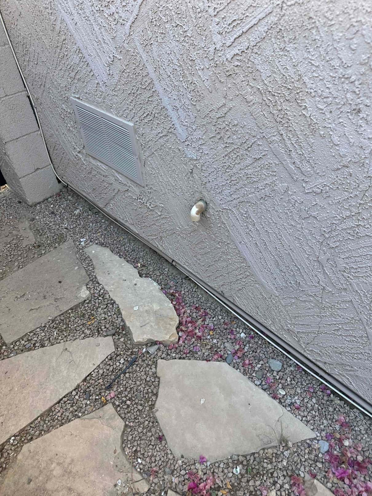 PVC drain outlet protrudes from exterior stucco wall below a rectangular vent grille near ground level.