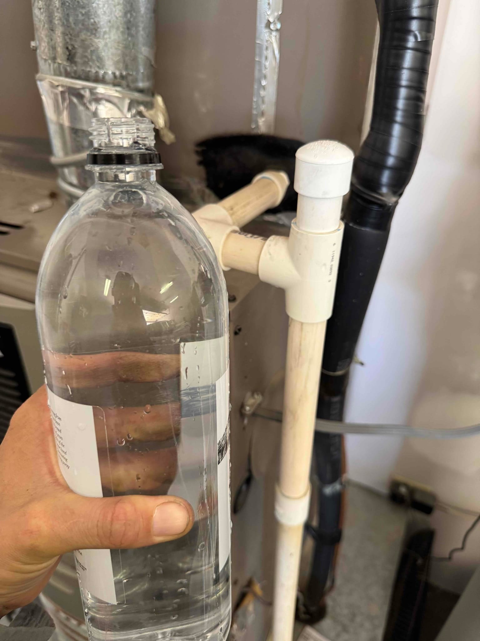 Condensate drain cleanout and PVC piping beside insulated refrigerant line at air handler, technician holding water bottle nearby.