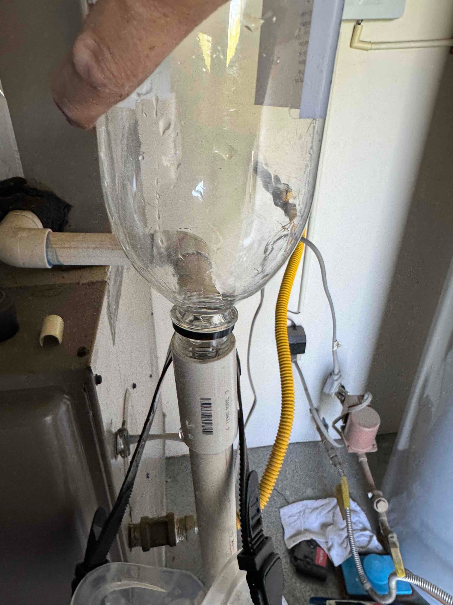 Hand holds a clear plastic bottle over a vertical PVC drain pipe near an indoor HVAC unit.