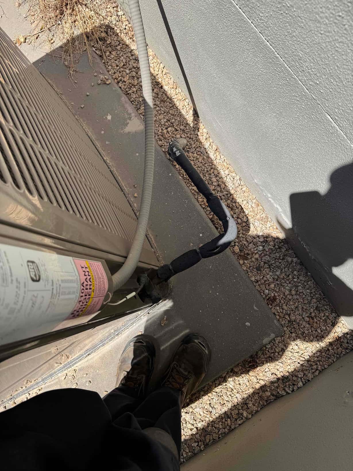 Insulated refrigerant line and electrical whip entering outdoor condenser at base beside technician’s boots.