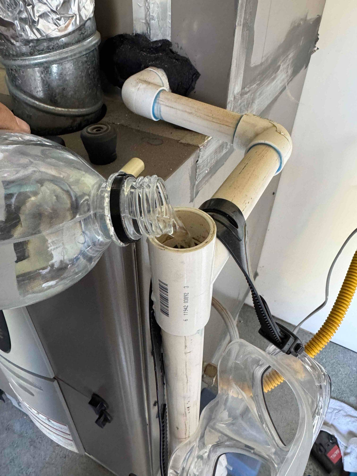 Liquid is poured into the PVC condensate drain cleanout at the furnace, with surrounding duct and piping visible.