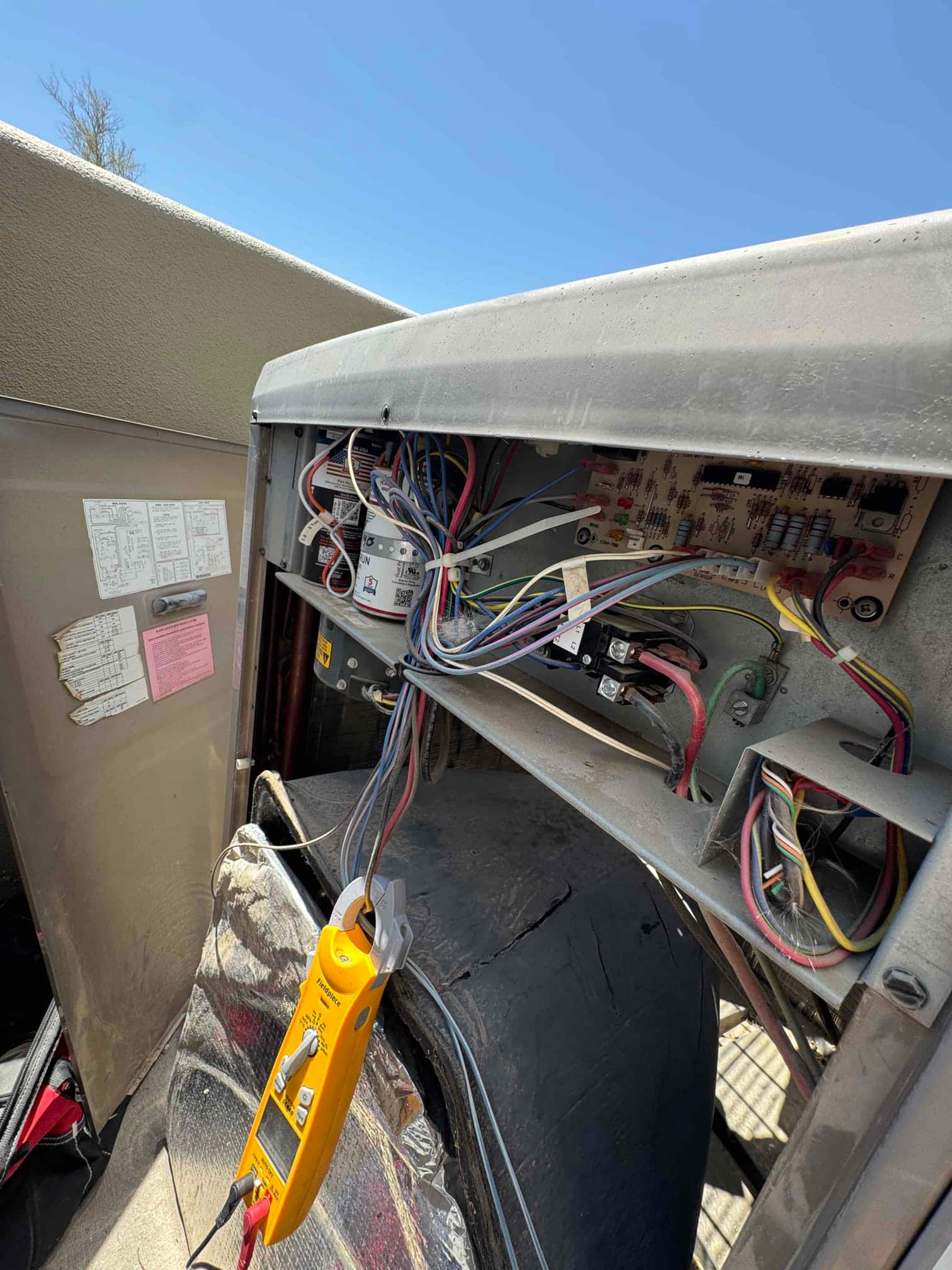 Open condenser control panel shows wiring, a dual-run capacitor, contactor, and control board, with a clamp meter attached to a conductor.