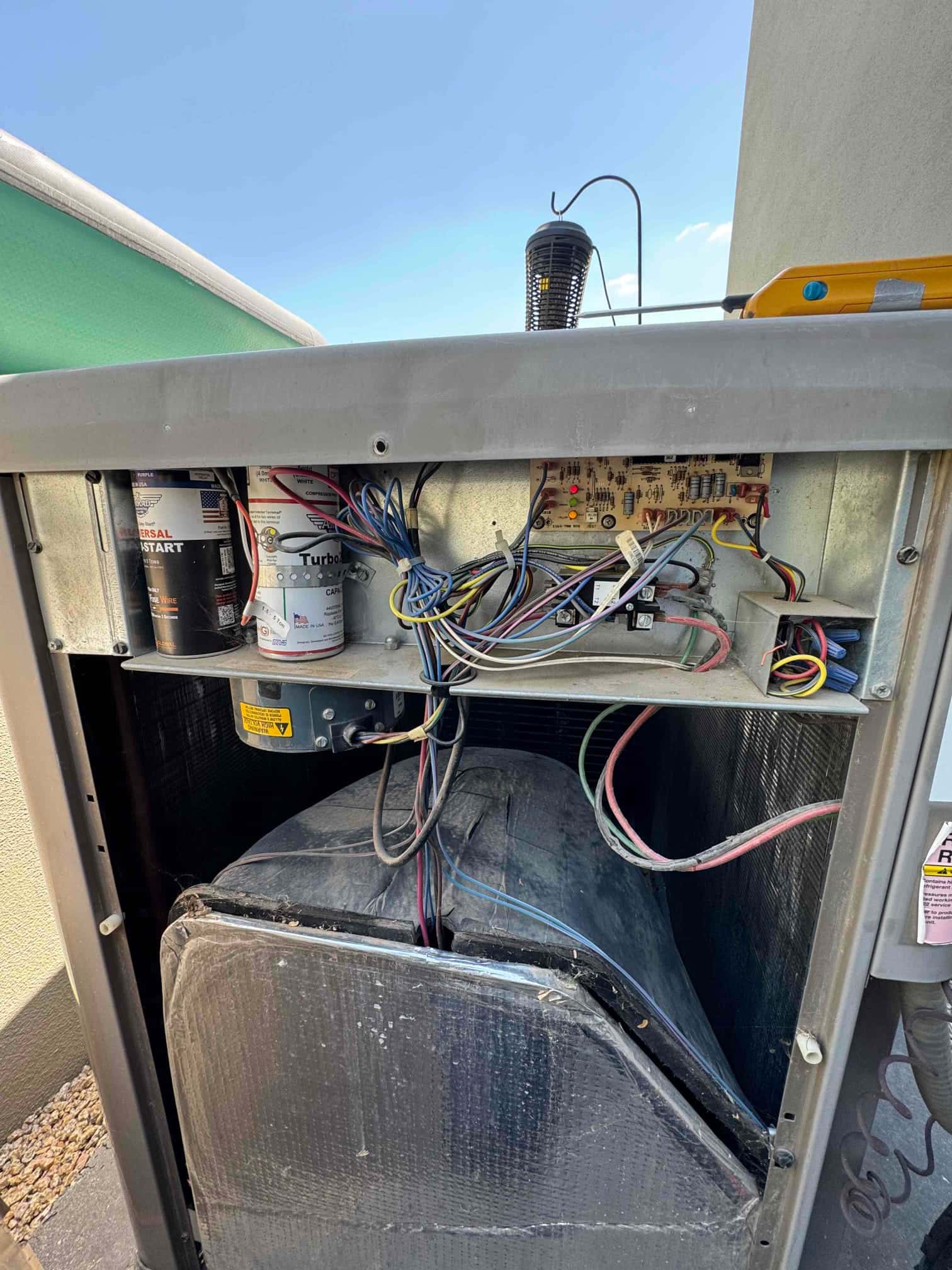 Open HVAC unit cabinet showing control board with wiring, dual-run capacitor, hard-start kit, and insulated blower housing exposed.