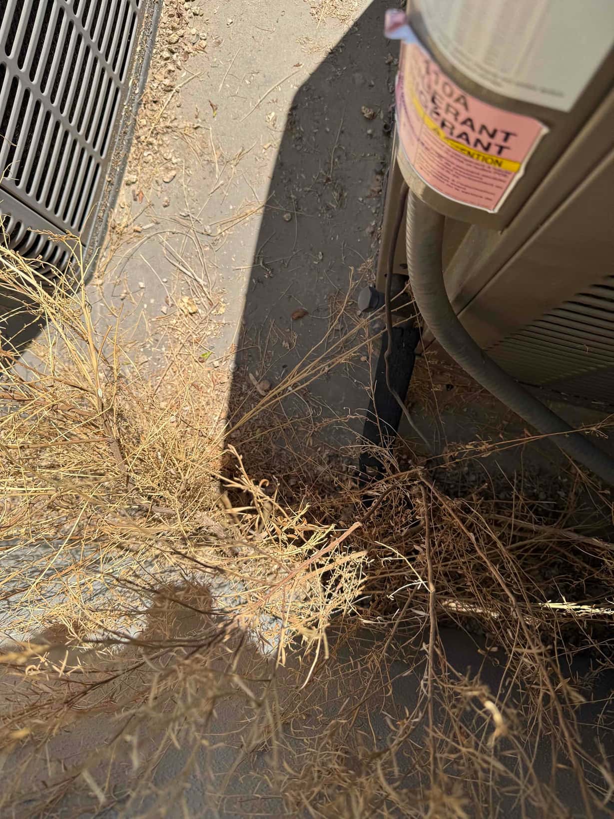 Outdoor condenser beside another unit with dry weeds and debris around the base and refrigerant line entering cabinet.