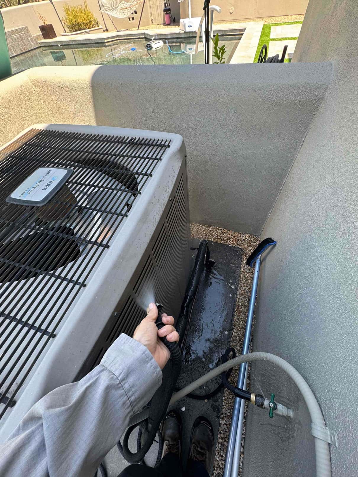 Outdoor condenser unit as a technician rinses the condenser coil with a hose sprayer; water drains below.