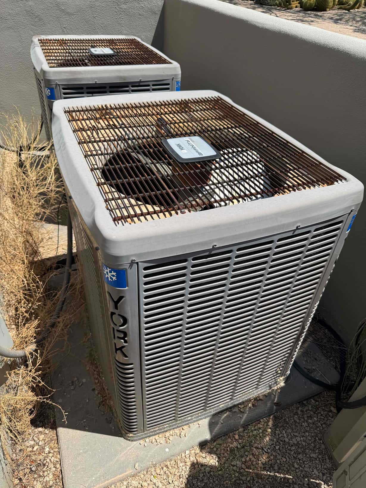 Two outdoor condenser units beside a wall; fan guards show heavy rust on the top surfaces.
