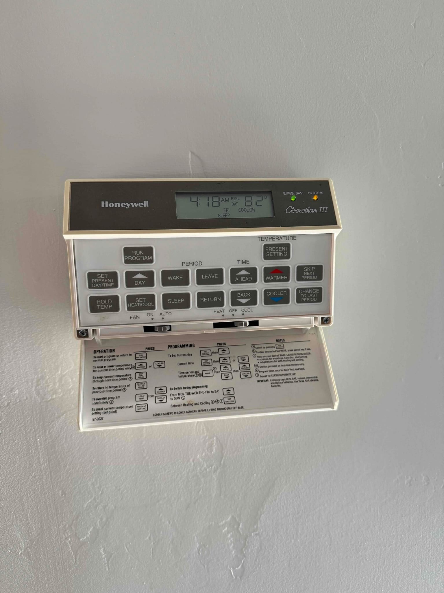 Programmable wall thermostat with open instruction cover, display indicates 82°F and 'COOL ON', with fan and system selector switches visible.
