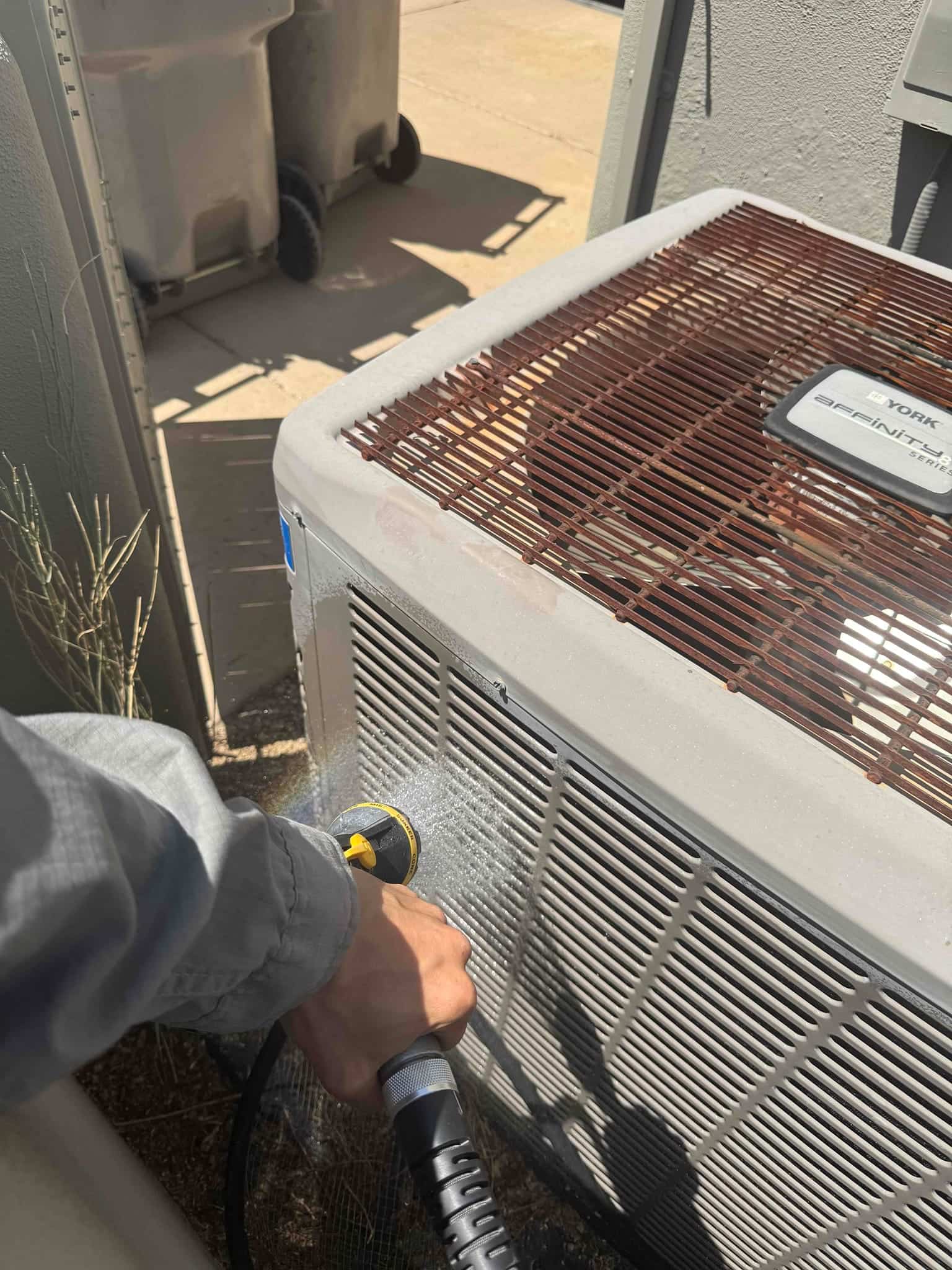 Technician sprays water onto the outdoor condenser coil fins with a hose nozzle.