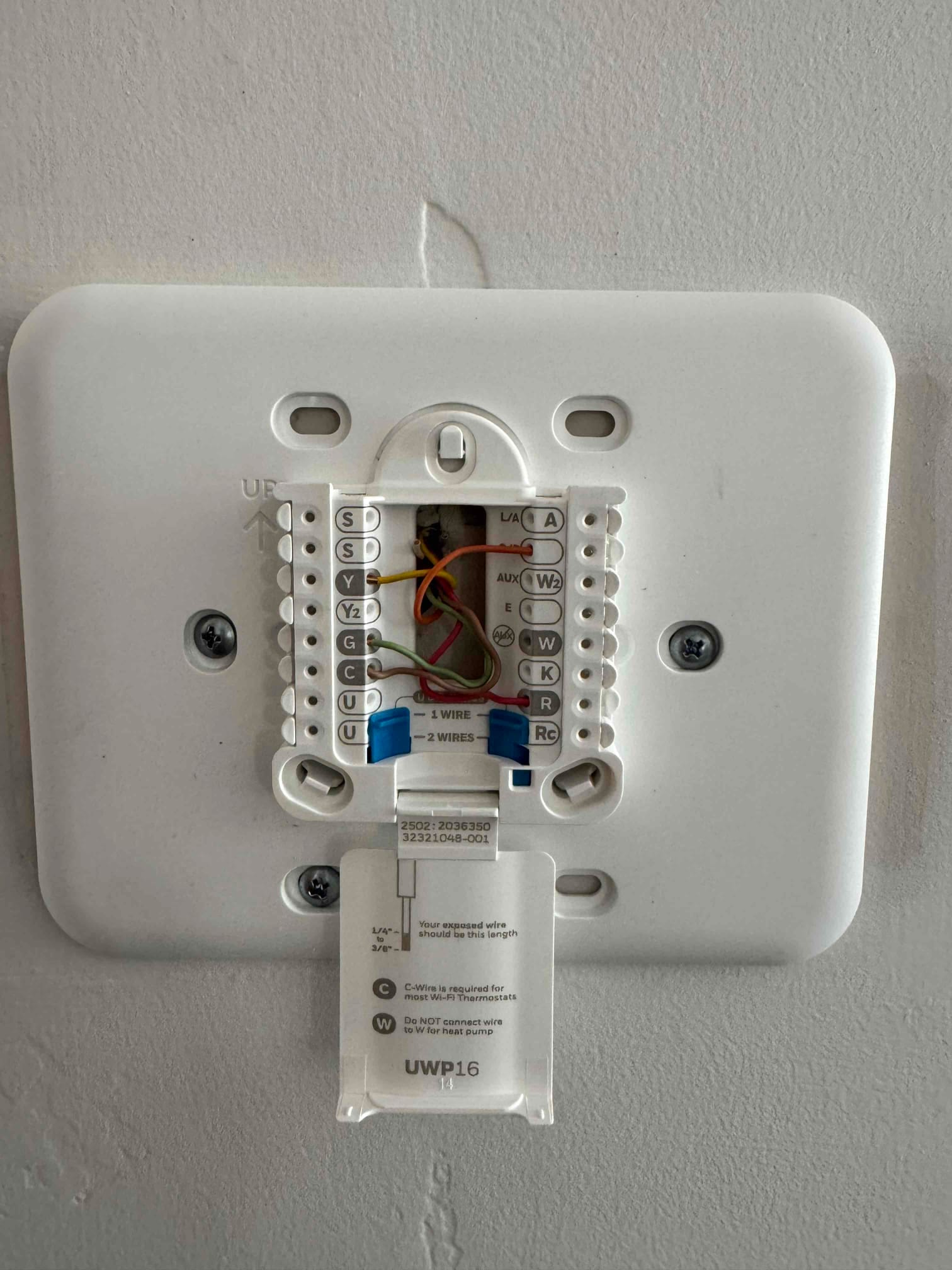 Thermostat wall plate with multiple low-voltage wires connected to labeled terminals, with mounting screws and the installation guide flap open.