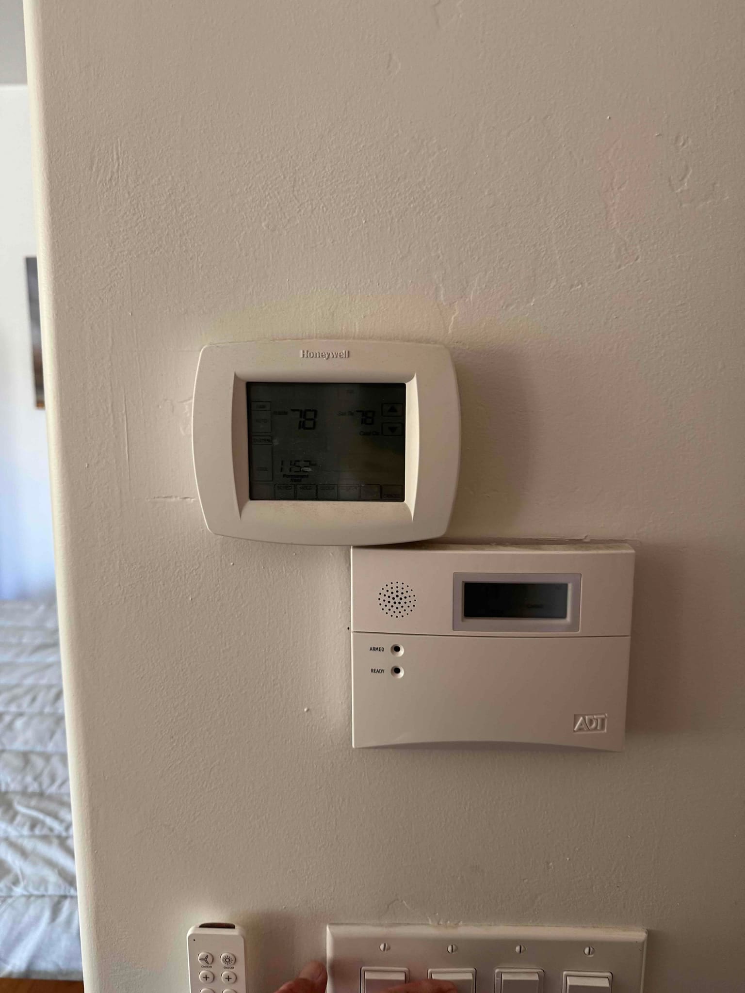 Wall-mounted digital thermostat above a home alarm panel, screen illuminated with temperature and control icons.