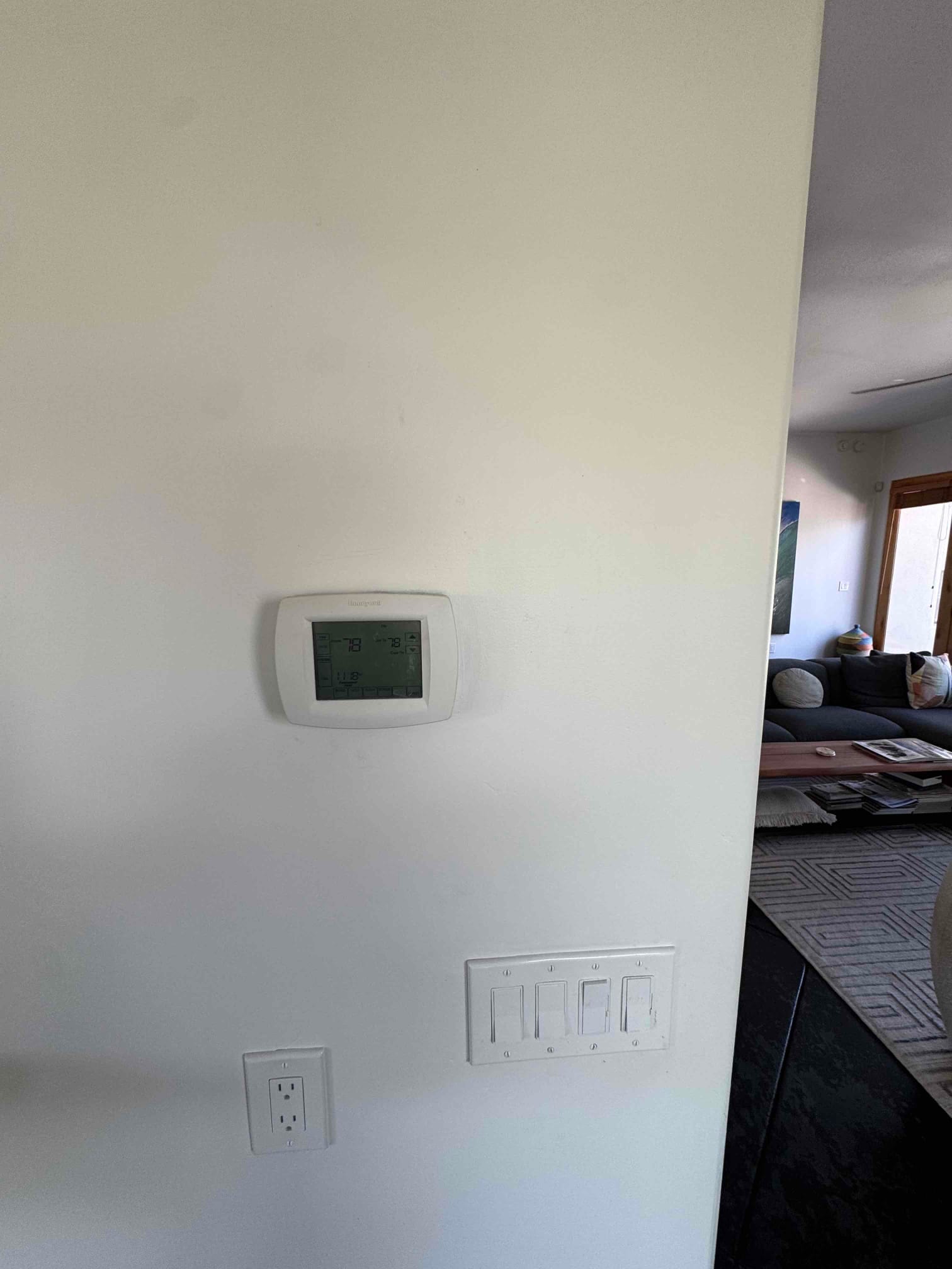 Wall-mounted thermostat with backlit display shows cooling mode, current room temperature, and setpoint in a living area.