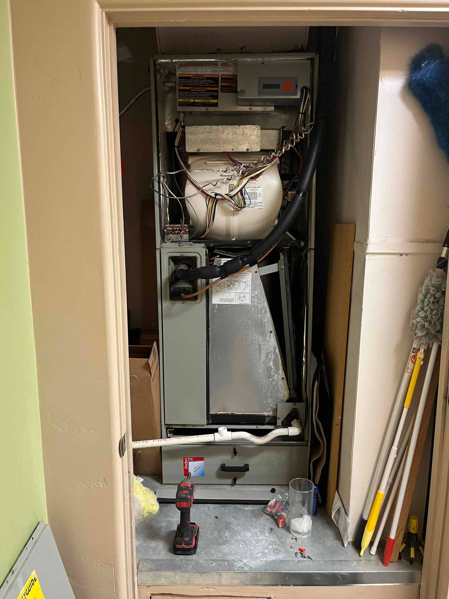 Air handler with access panels removed, showing blower housing, control wiring, and condensate drain assembly inside a closet.