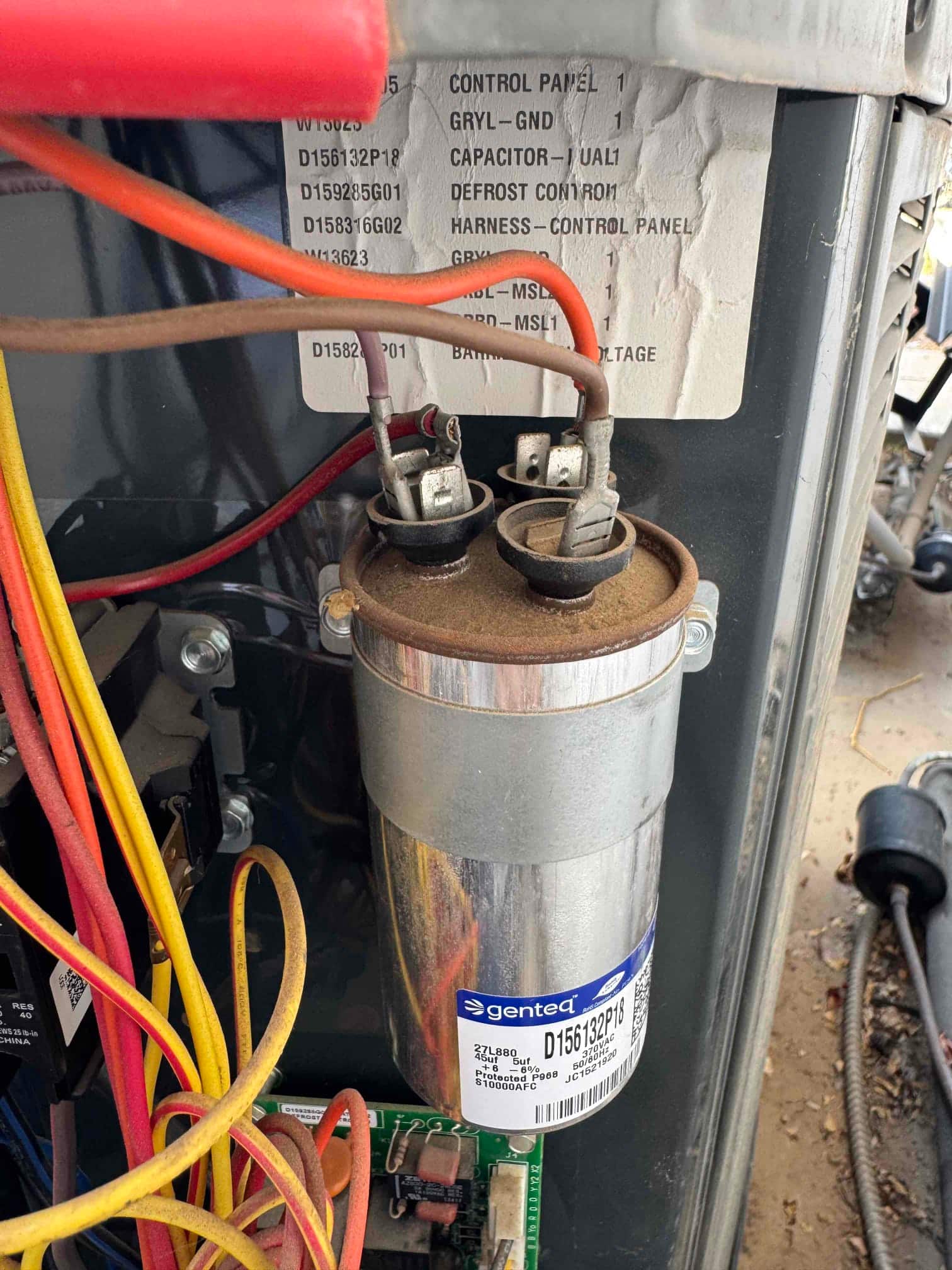 Dual-run capacitor mounted in the outdoor unit control compartment with wires attached to its terminals and dust on top.