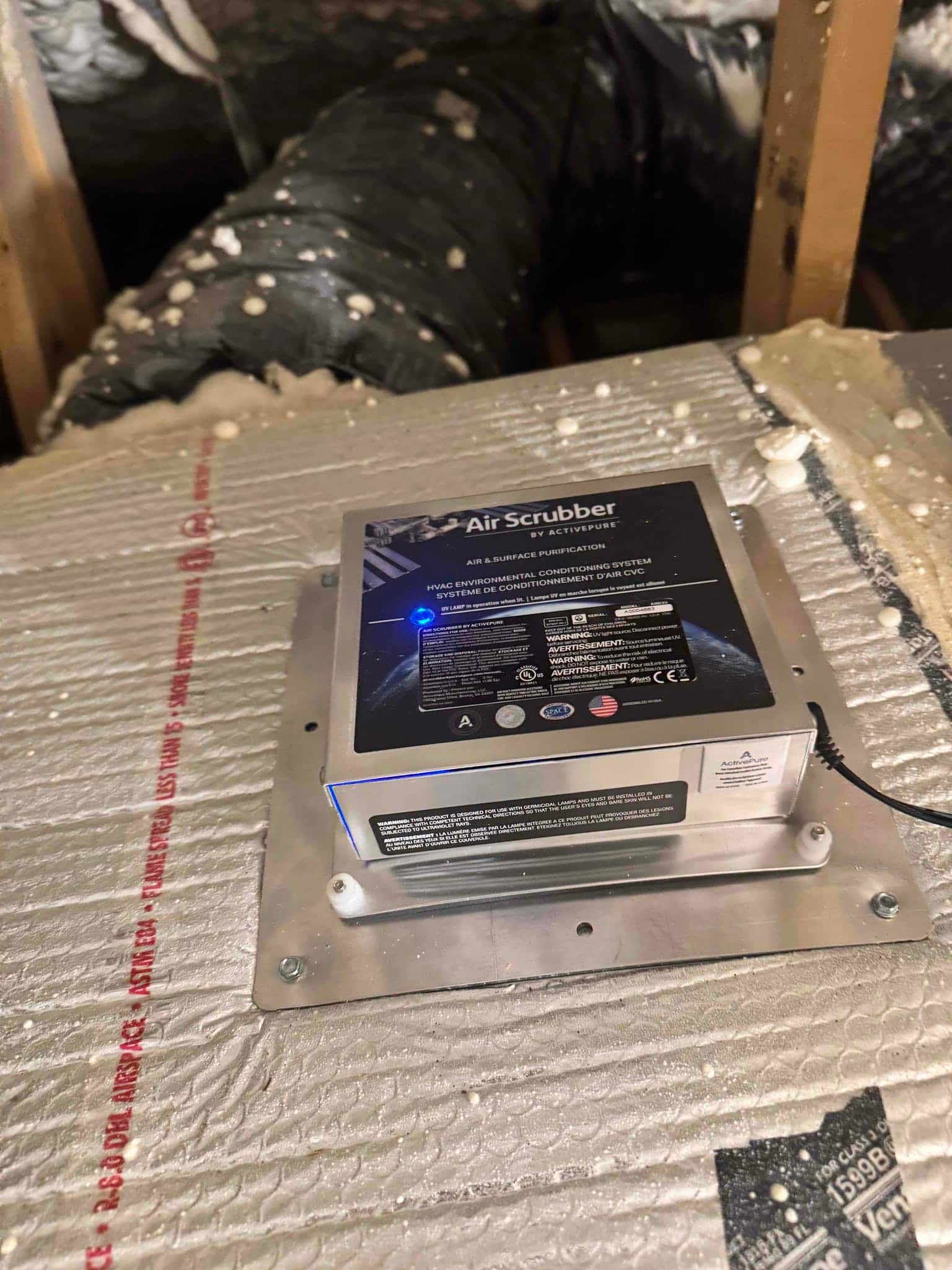 In-duct air purification device mounted to ductboard plenum with blue indicator light illuminated, with flexible duct and spray foam insulation visible.