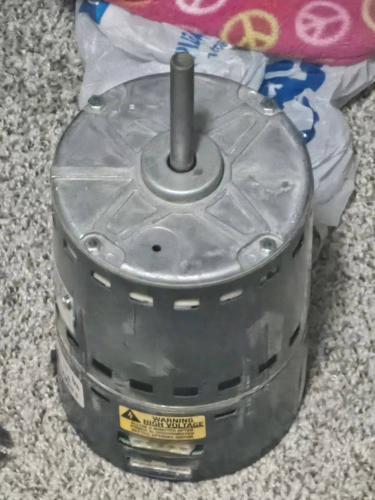 Electric HVAC fan motor removed from unit, showing top shaft and vented housing with high-voltage warning label.