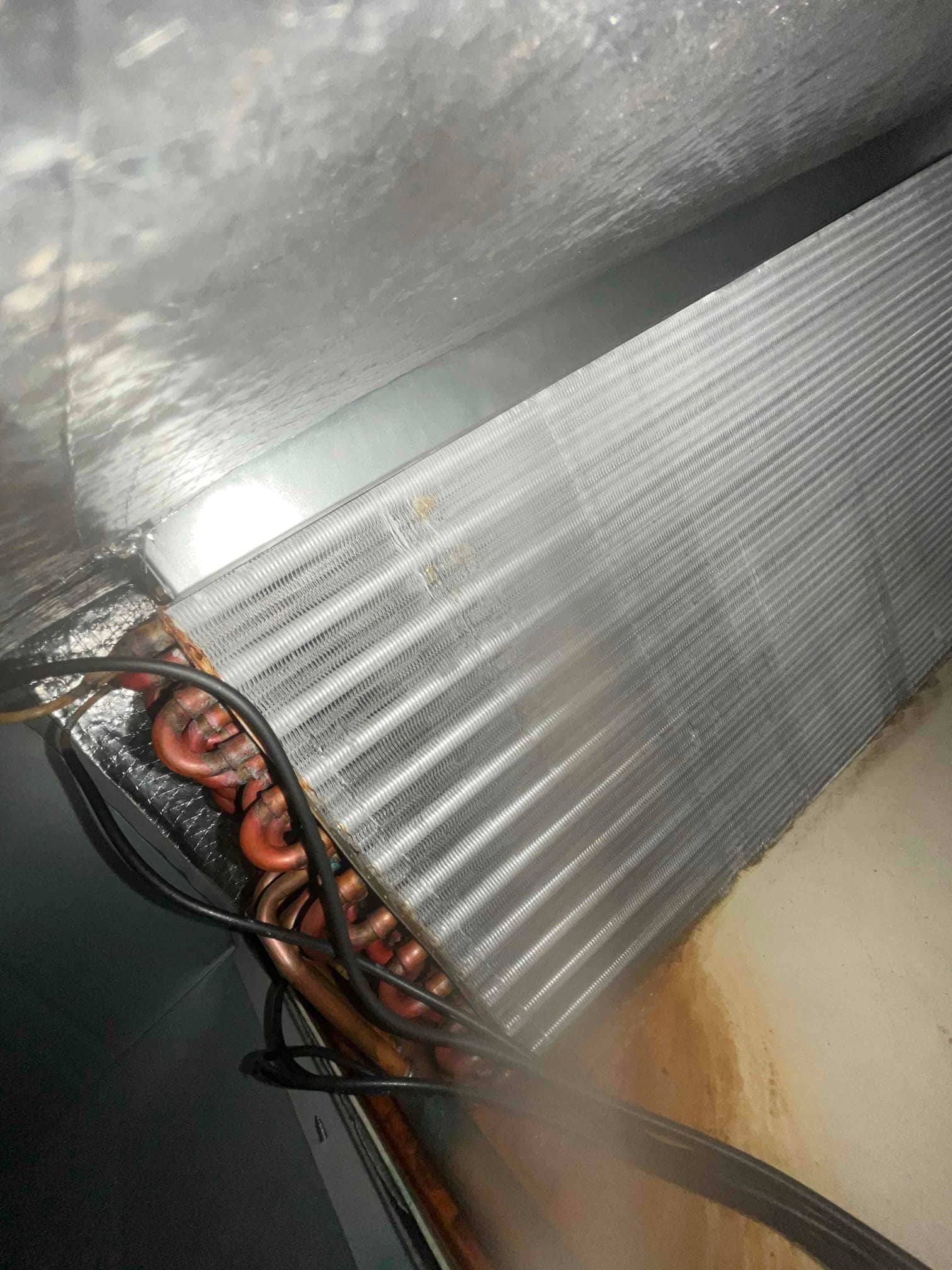 Evaporator coil with copper tubing and aluminum fins above a drain pan showing rust-colored staining.