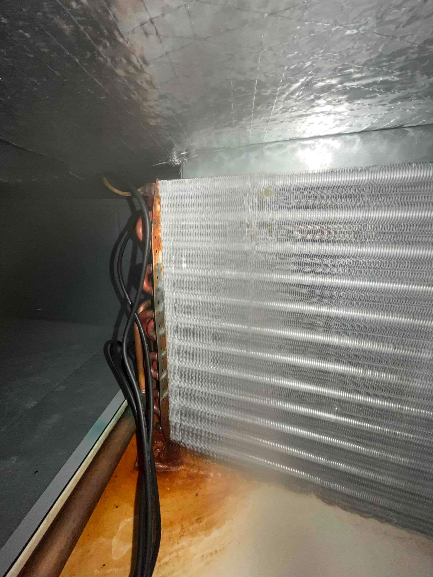 Evaporator coil inside air handler with copper tubing on the side and rust-stained drain pan below.