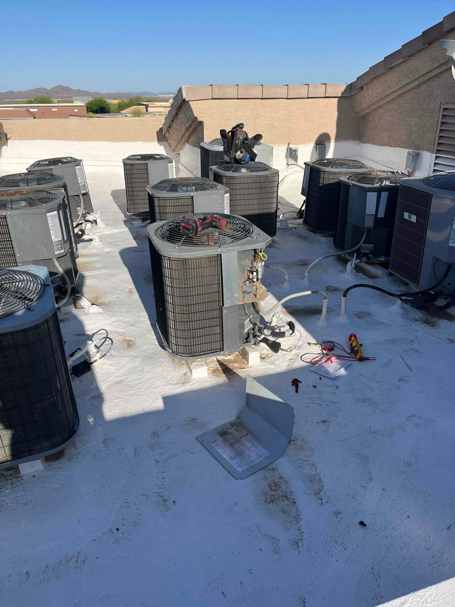 Multiple rooftop condenser units; one cabinet panel removed, exposing wiring components, with tools and a multimeter on the roof.