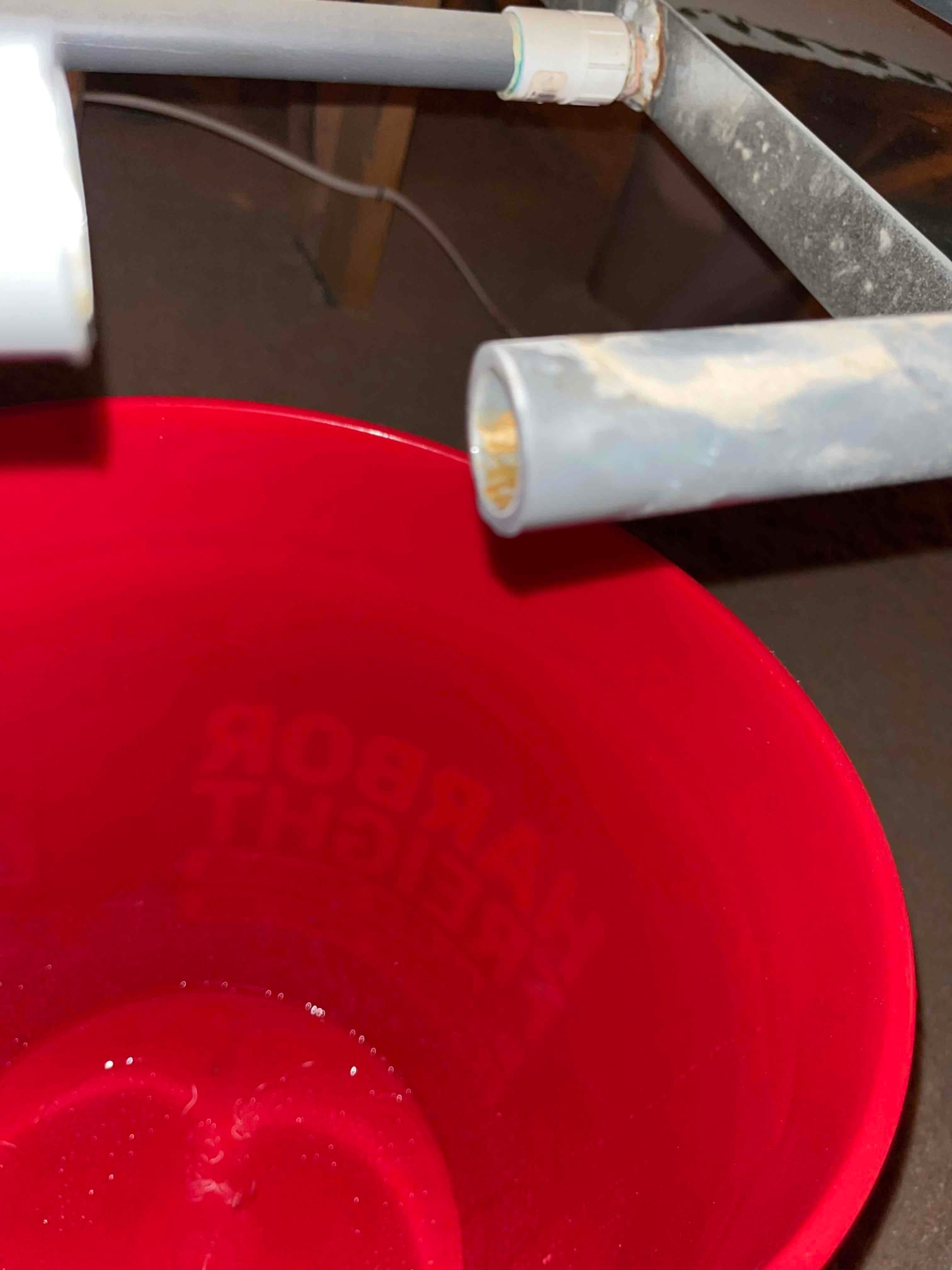 Open PVC condensate drain pipe positioned over a red bucket beside a dusty drain pan.