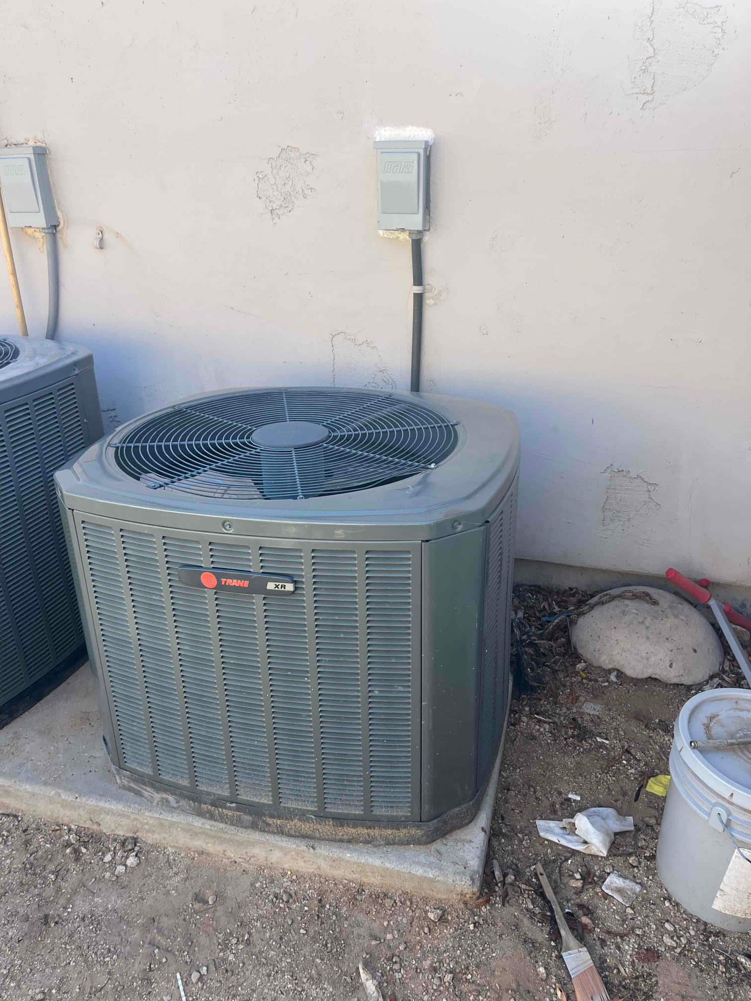 Outdoor air-conditioning condenser unit on pad beside wall, with disconnect boxes and surrounding ground debris.