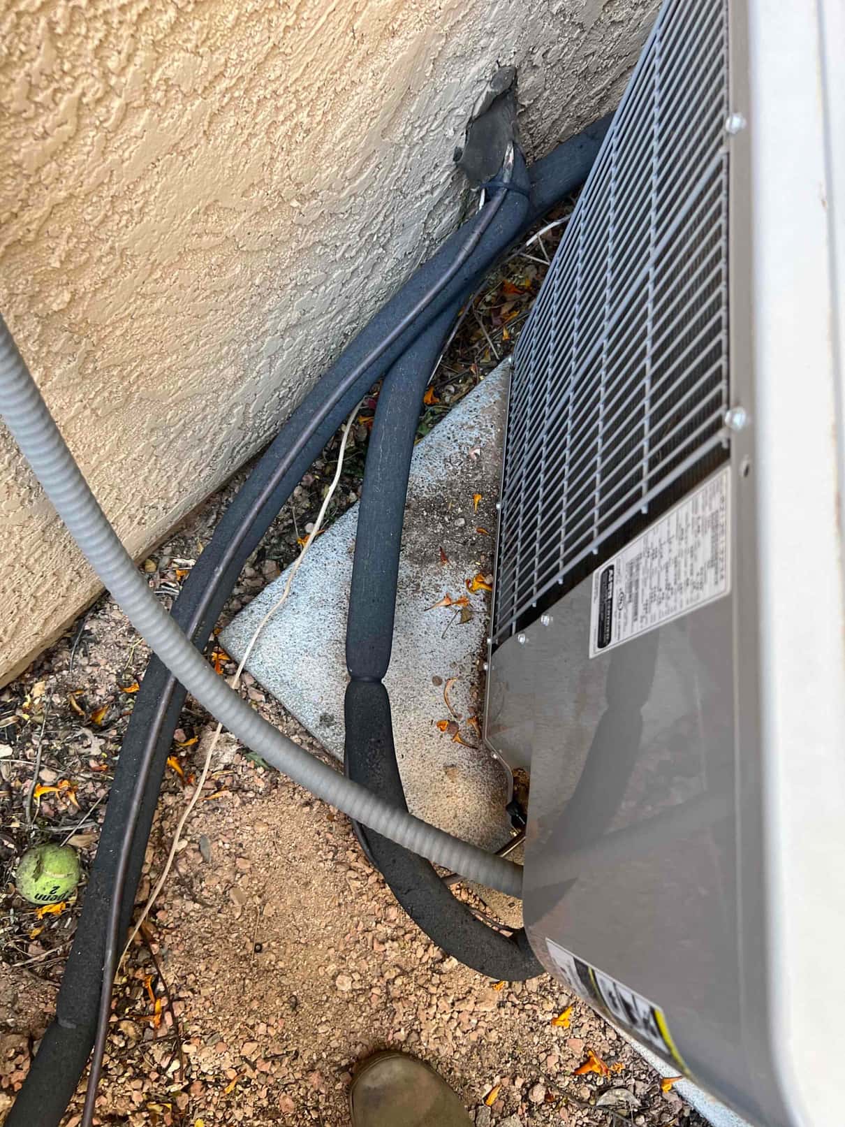 Outdoor condenser beside stucco wall with insulated refrigerant lines and flexible electrical conduit entering the wall penetration.