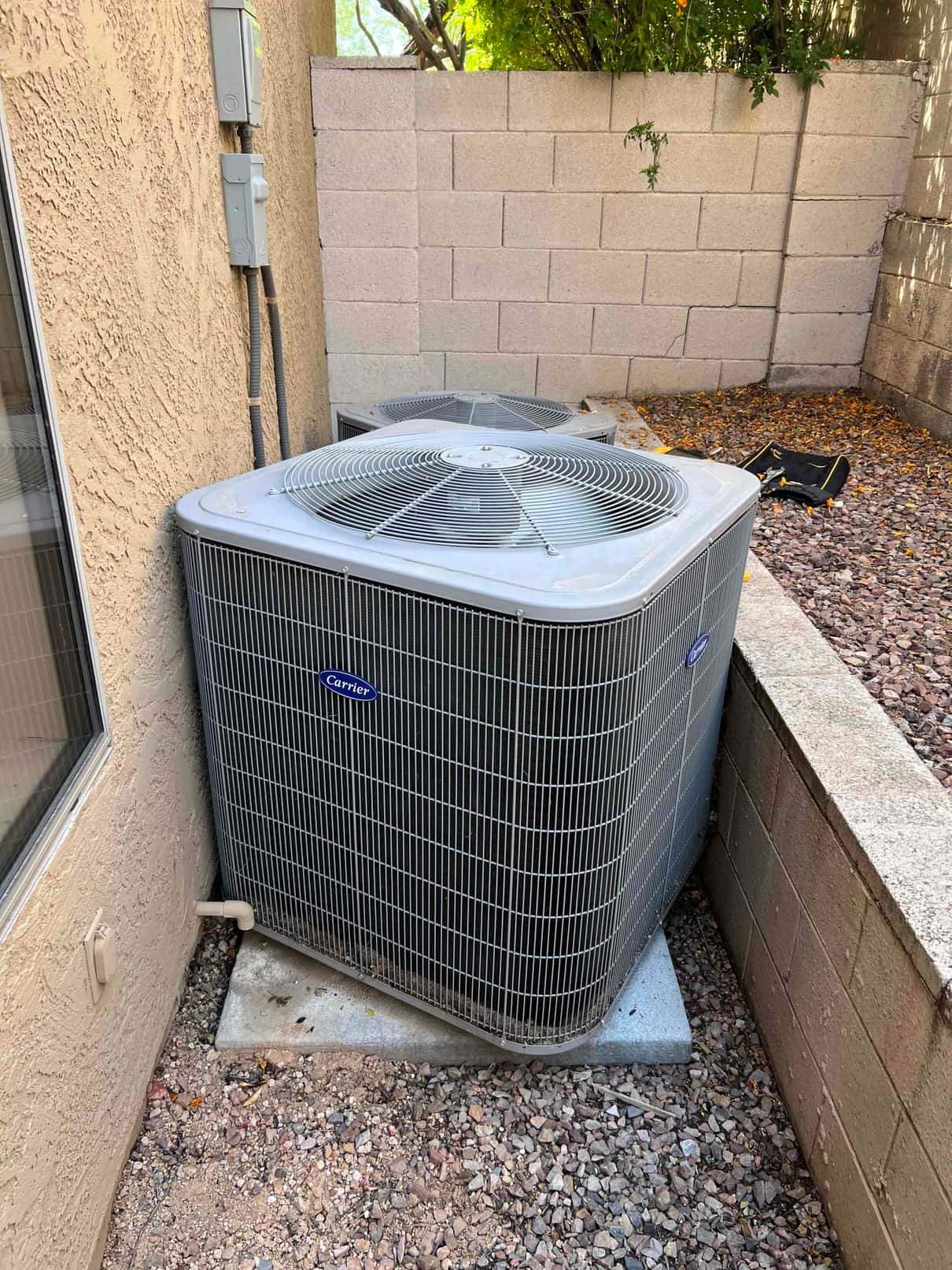 Two outdoor condenser units beside a stucco wall with electrical disconnects and refrigerant lines.